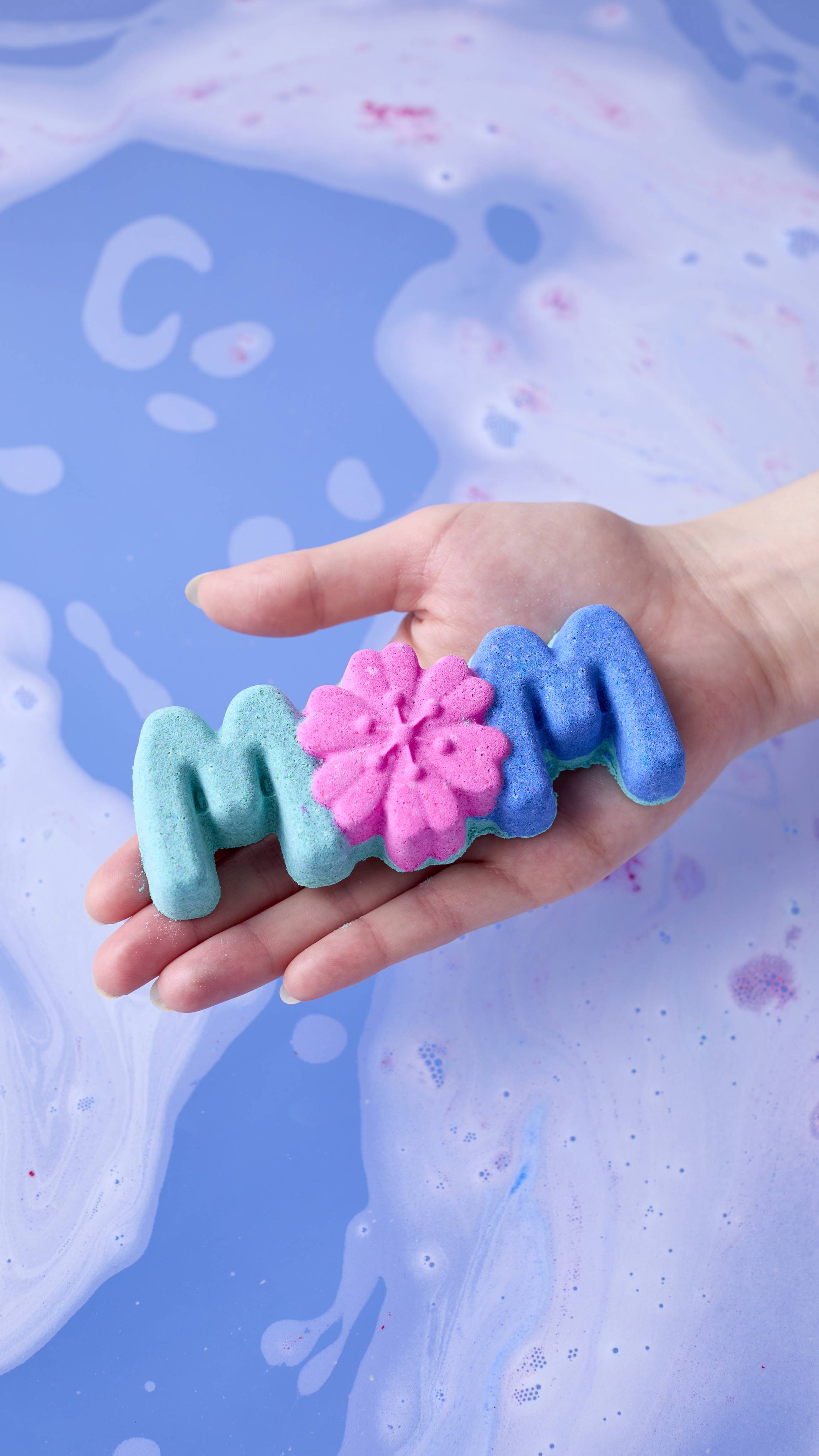 A hand holds the "Mum" bath bomb over milky blue bathwater. The product features a teal "M," a pink flower-shaped "O," and a dark blue "M," contrasting against the water's soft, cloudy texture.