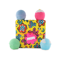 Mum. A square gift box wrapped in yellow paper designed with bold flowers in a hand-drawn style, tied with an orange string and pink tag. Four themed bath bombs sit around the box.