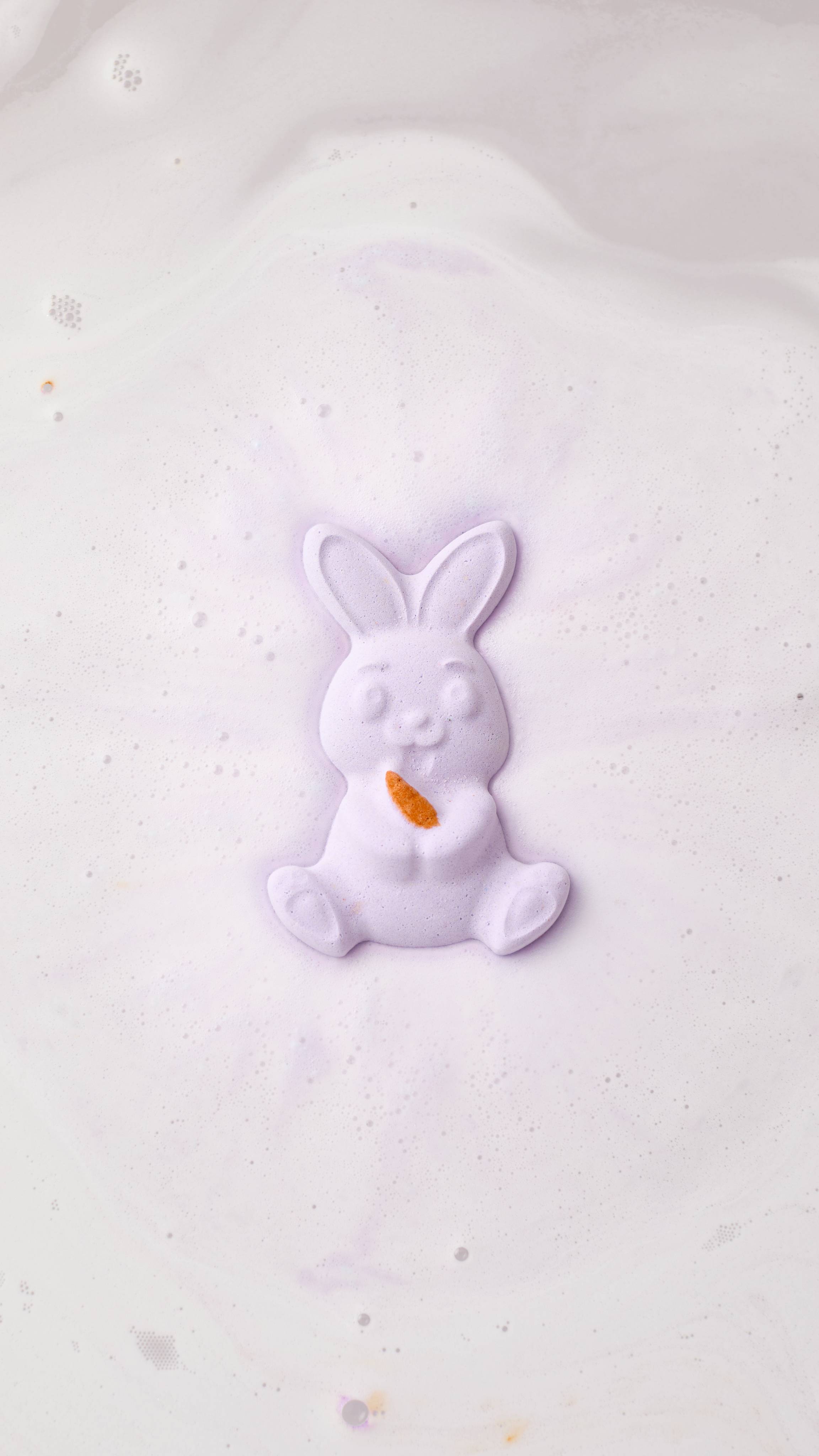 An aerial view image of the Lush "Munching Bunny" rabbit-shaped bath bomb, creating a swirl of pale purple foam. The bunny has a small orange carrot detail on its paws. The product dissolves, leaving a thick blanket of velvety lilac foam. 