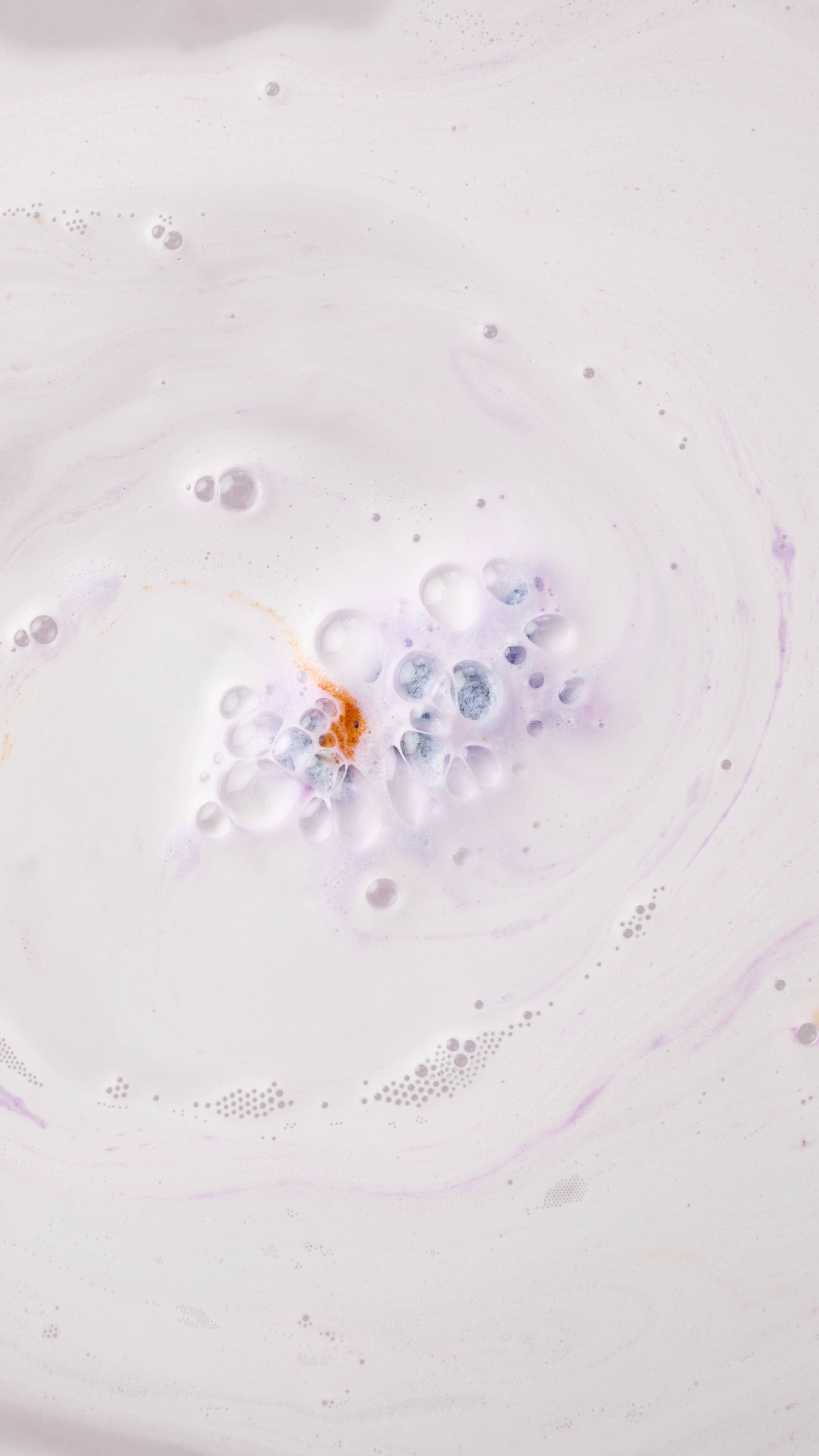 The Munching Bunny bath bomb dissolves in the center of a bath, creating a swirl of creamy white and pale purple bubbles. A small concentrated patch of orange and blue foam emerges from the center of the fizz.