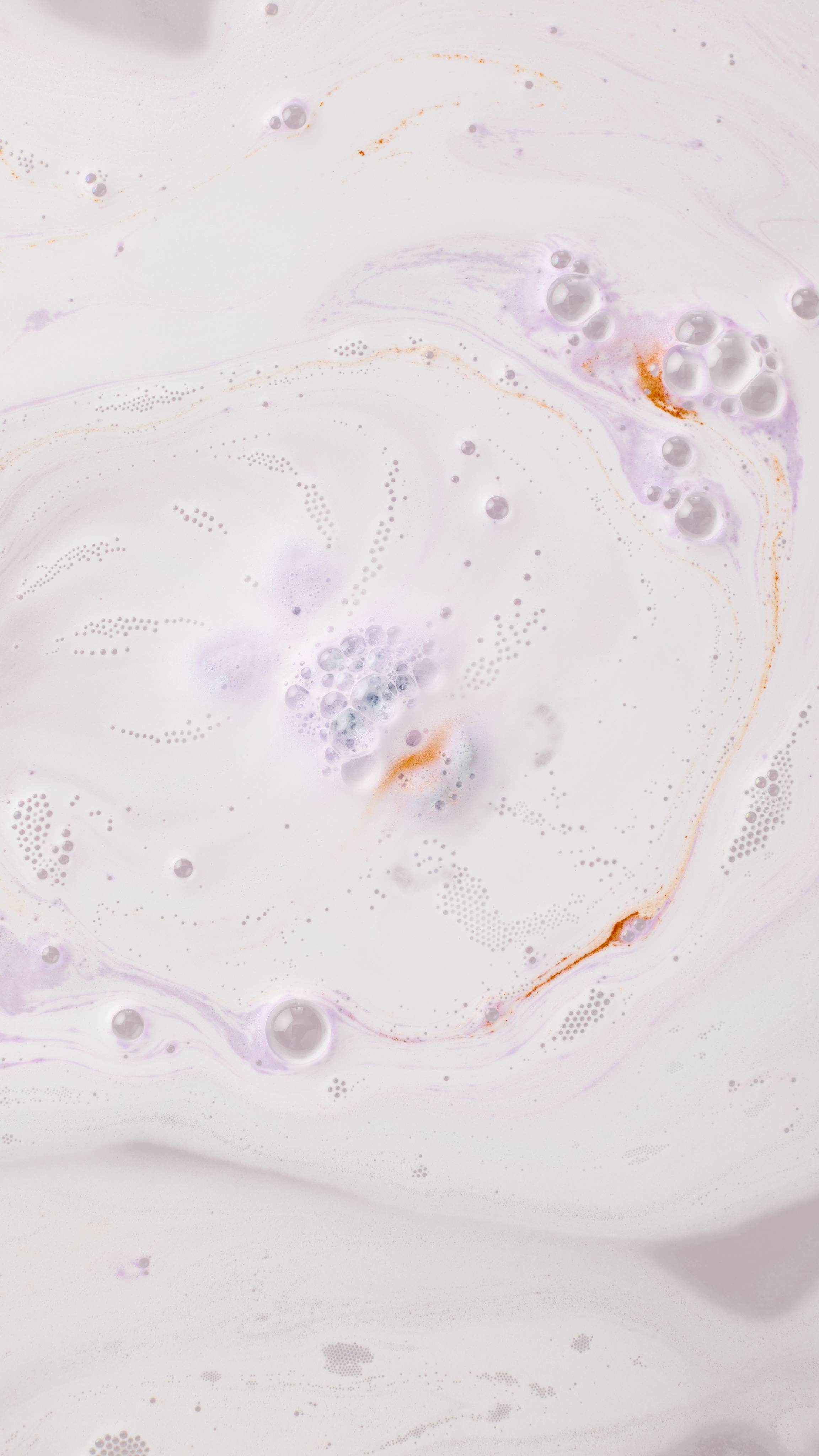 The Munching Bunny bath bomb dissolves into creamy, milky white bathwater, releasing soft trails of lavender-purple and delicate swirls of orange. Small clusters of bubbles pop on the surface as the colors gently blend.