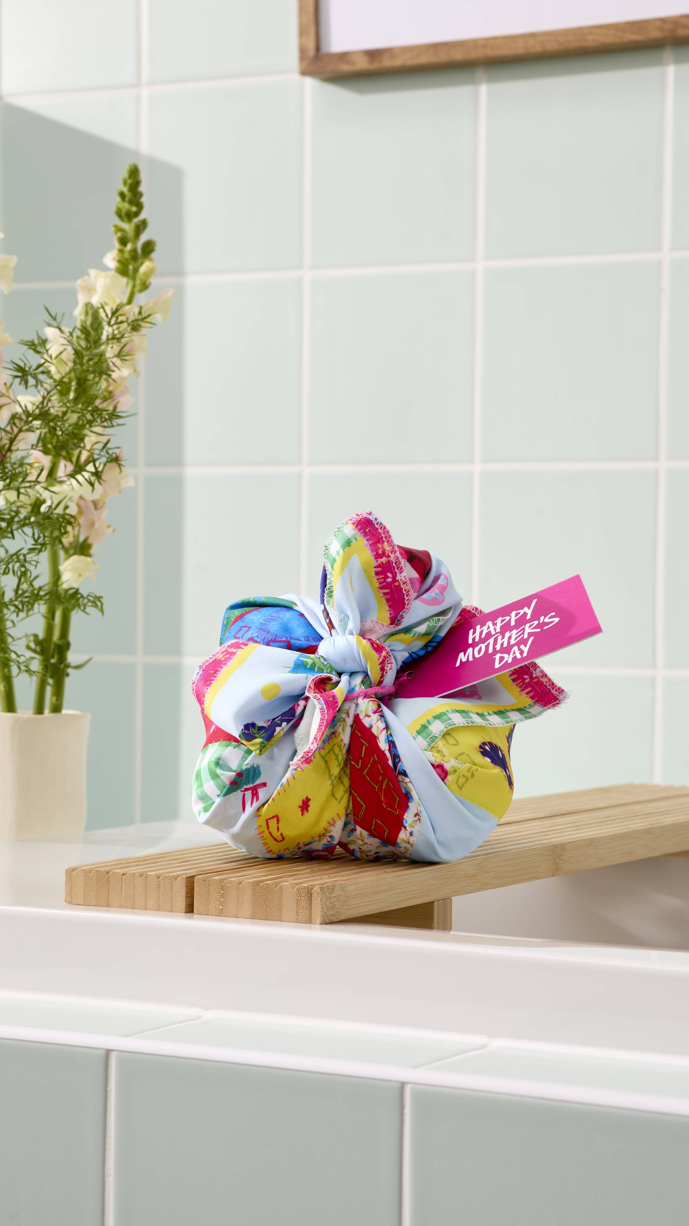 The image shows the "My Family Houses" fabric knot-wrap featuring a playful house design. Tied into a decorative knot with a pink "Happy Mother's Day" tag, it sits on a wooden bath bridge against a background of pale green tiles and fresh flowers.