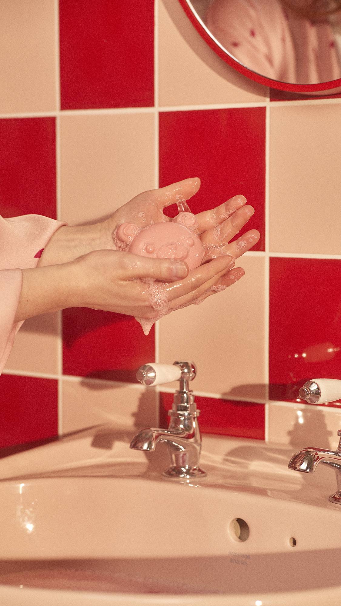 A close-up image of a sink in front of a red and pale pink checkered tile. The model is using the My Li'l Chia Bear soap. 