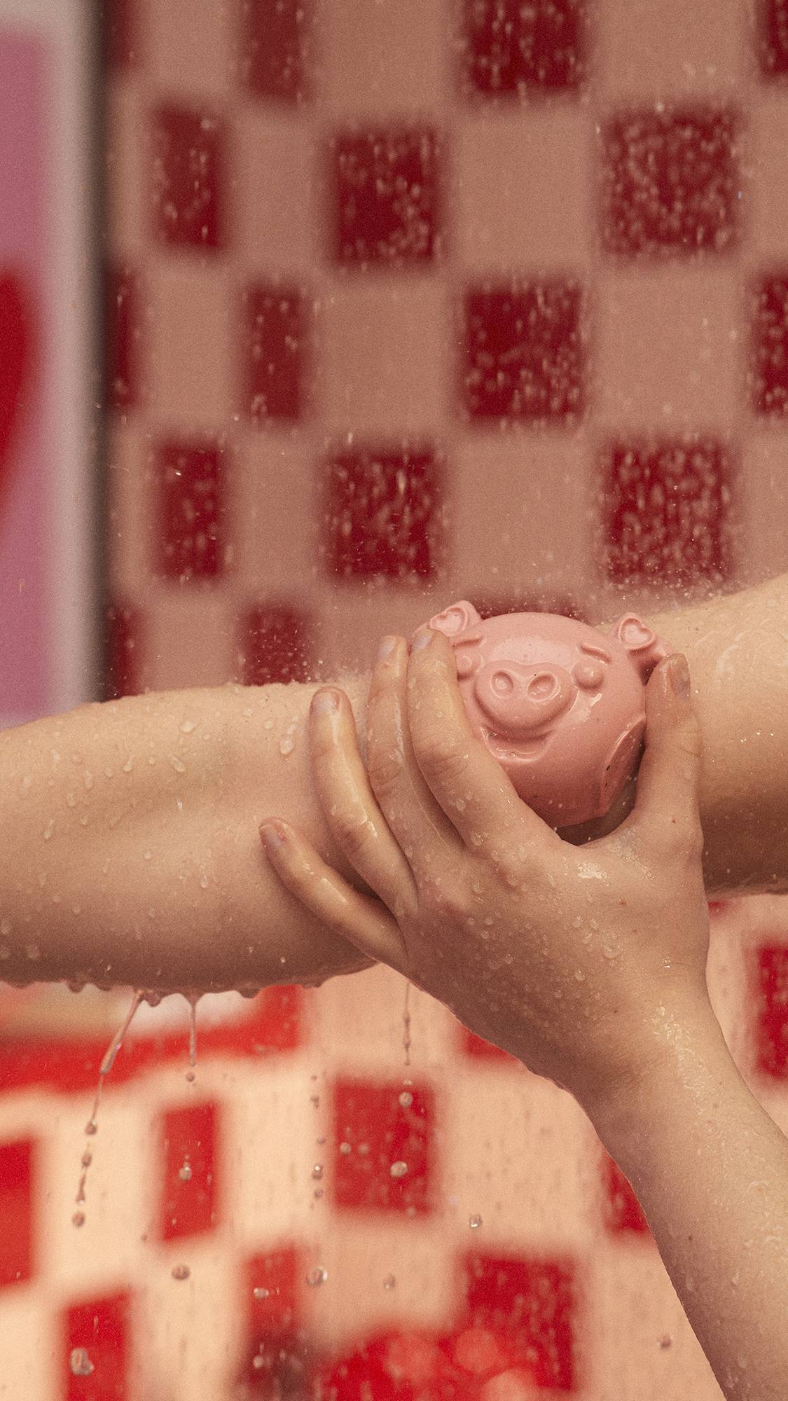 The image shows a close-up of the model lathering up their forearm with the My Li'l Chia Piglet soap under a running shower. 