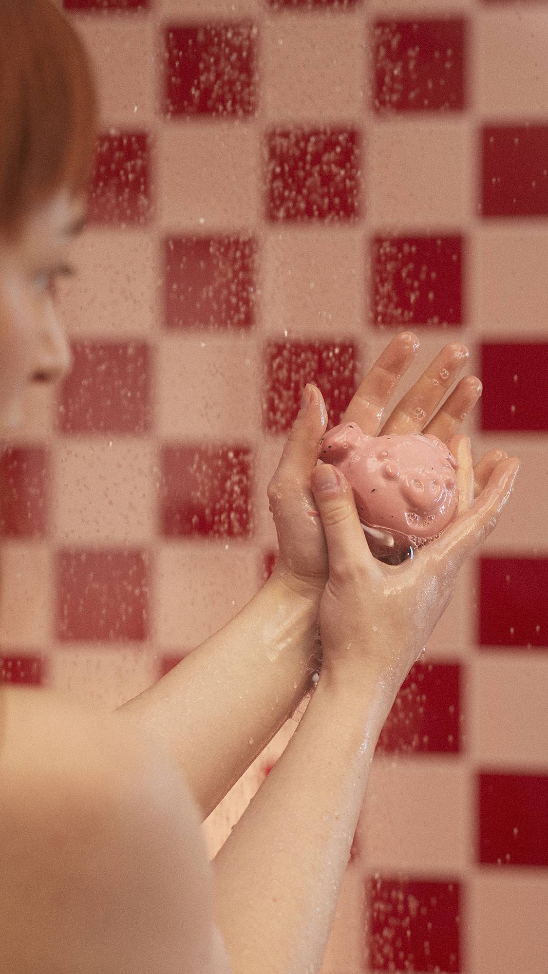 The image shows a close-up of the model's hands as they lather up soapy suds with the My Li'l Chis Piglet soap under running water. 