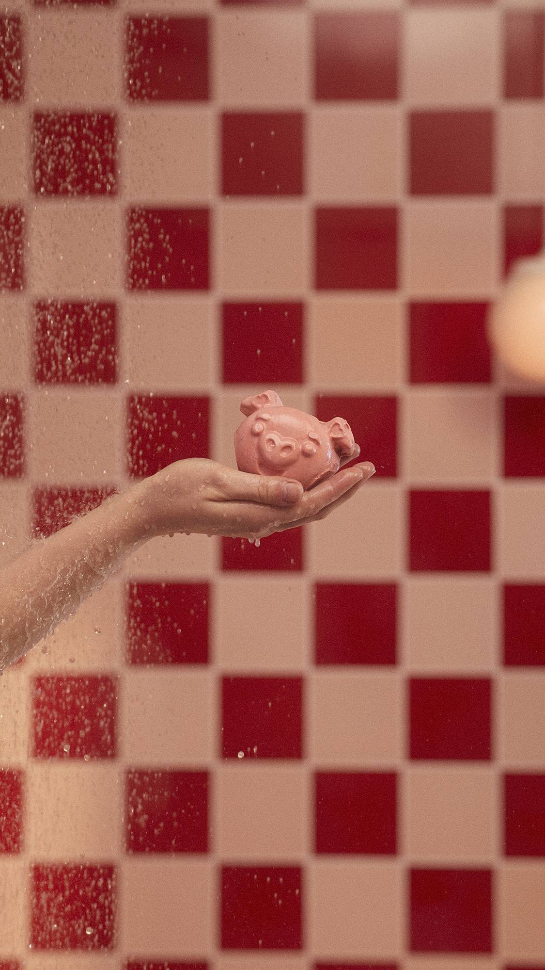 The image shows a close-up of the model's hand as they hold the My Li'l Chis Piglet soap in their hand under running water. 