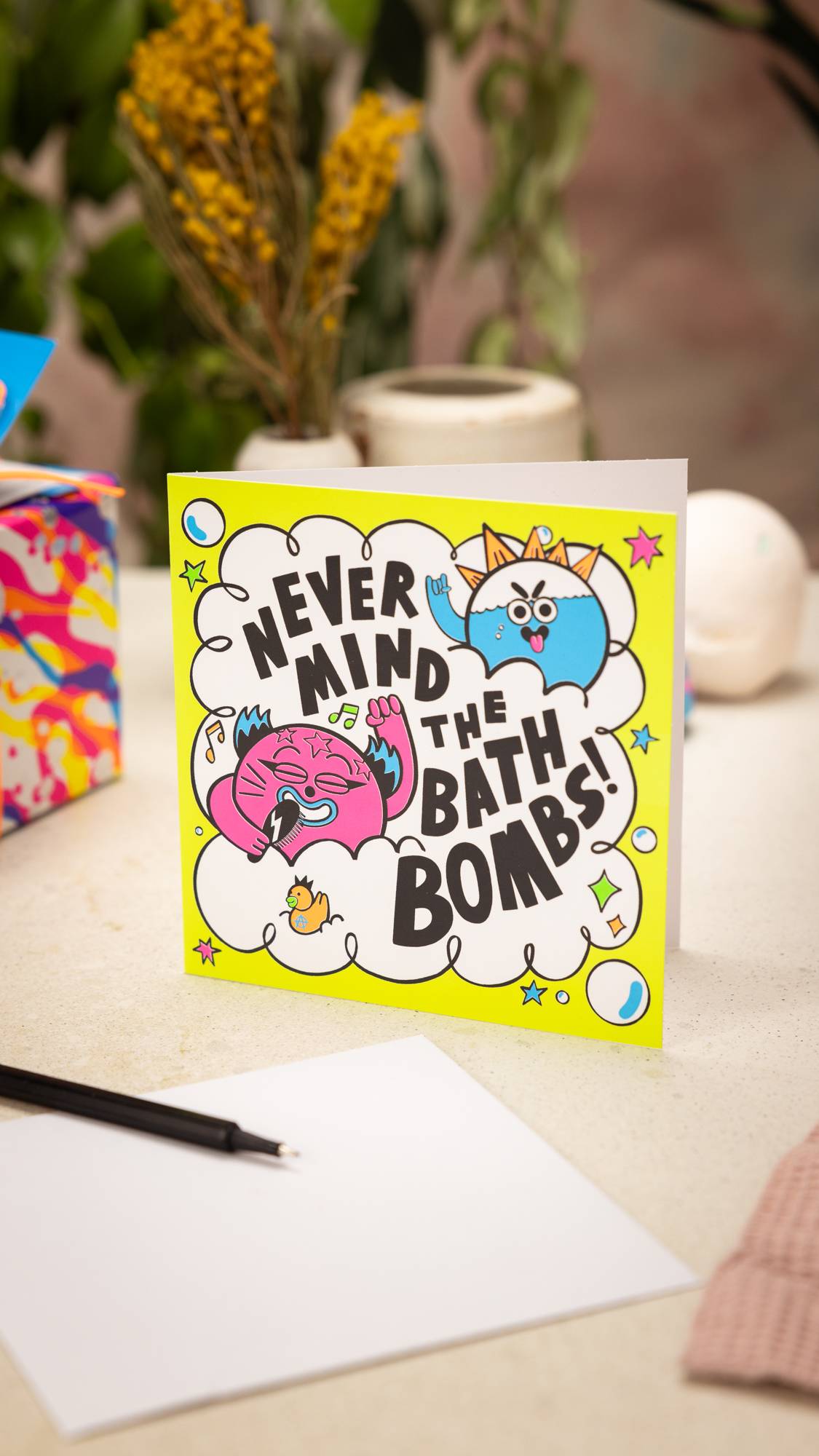 Image shows the Never Mind The Bath Bombs greetings card stood on a table. A pen and envelope are in front.