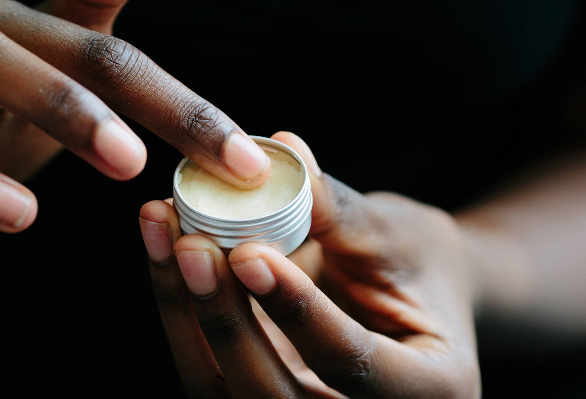 A finger is rubbed in a small round tin of thick, buttery yellow None Of Your Beeswax lip balm.
