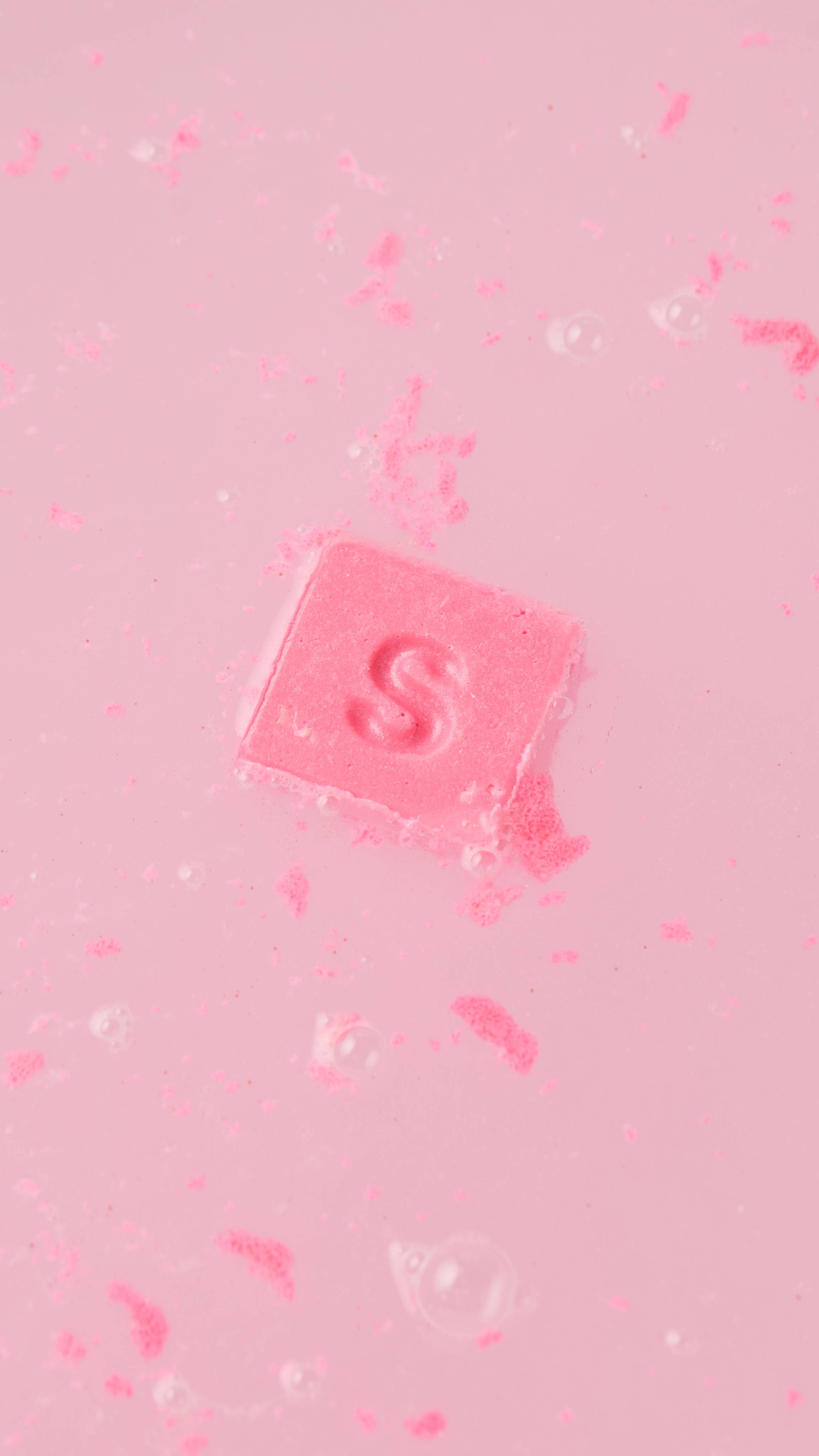 A solid, vibrant pink, square bath melt floats in a bright pink, bubbly water. The letter "S" is embossed on its surface, and tiny pink granules are scattered throughout the water.
