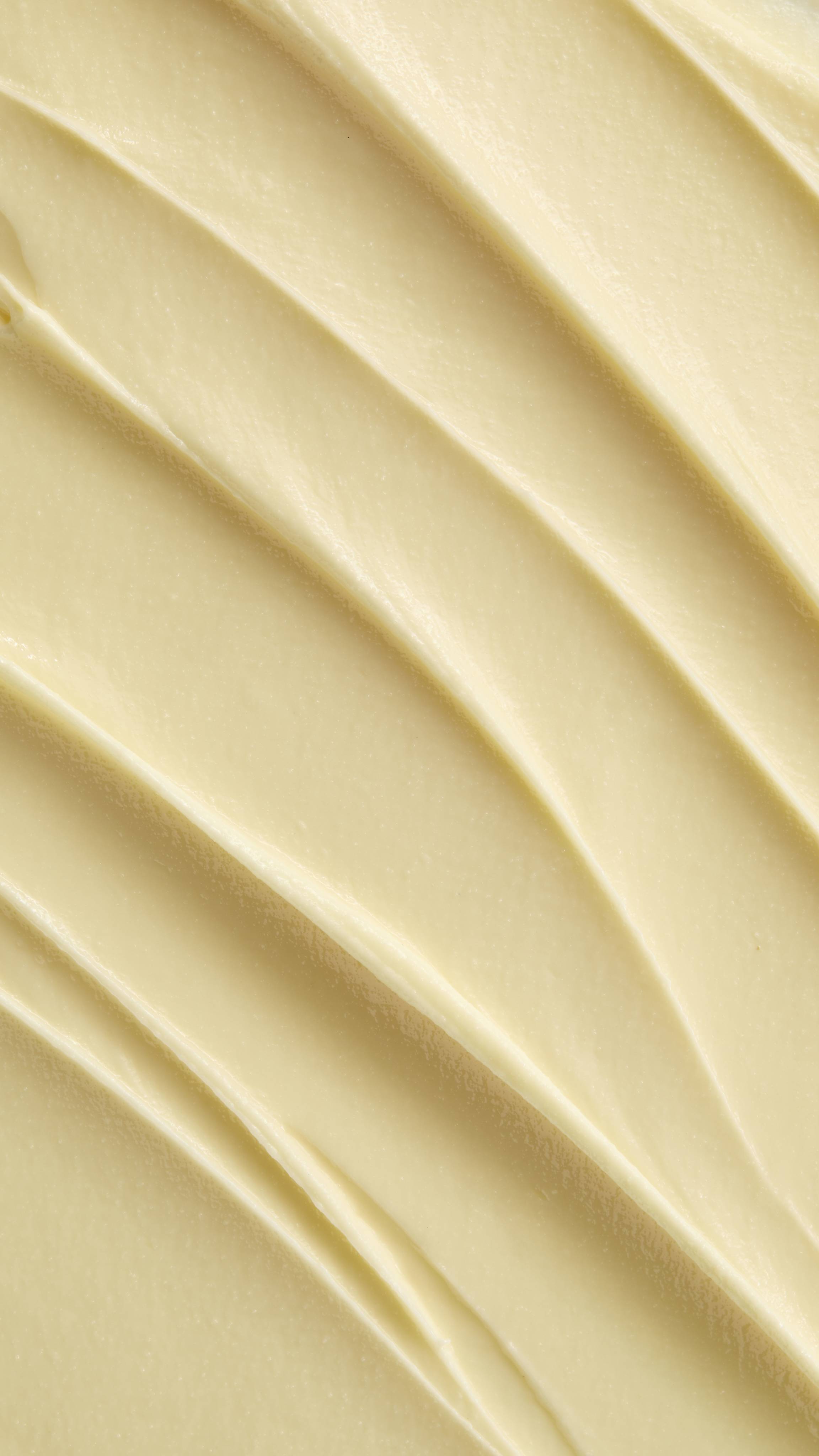 A super close-up image showing diagonal, overlapping smears of the thick, creamy, pale yellow Lush "Oat" facial cleanser. The smooth product has a matte finish and subtle ridged textures.