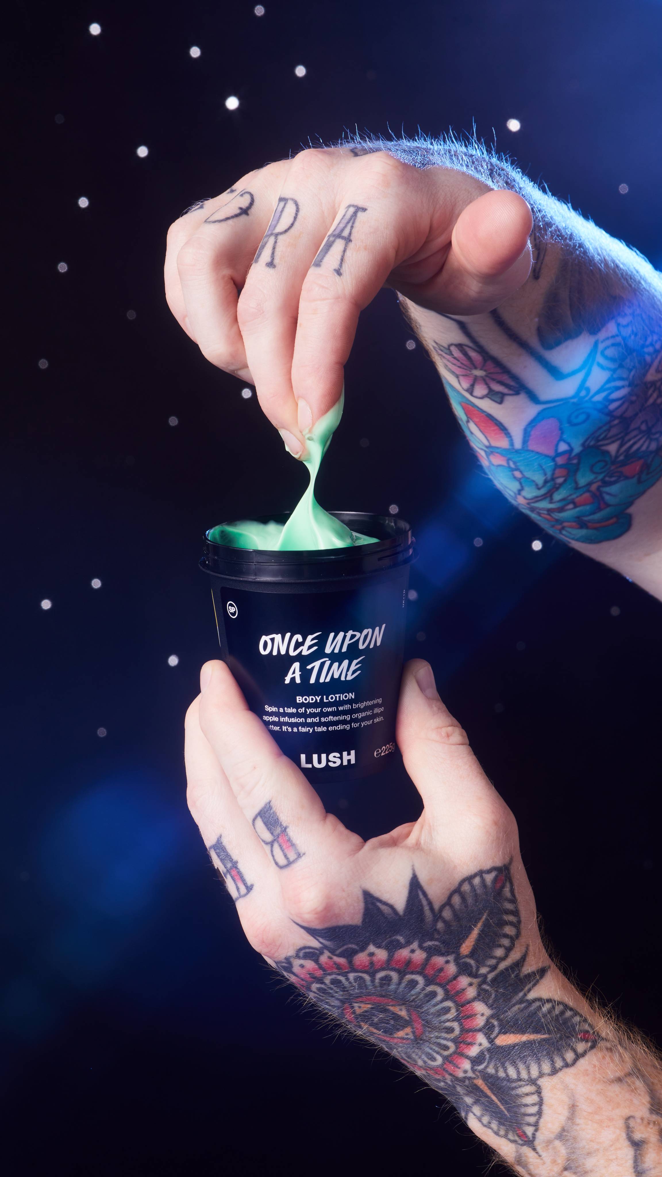 Model is holding the black LUSH pot of Once Upon A Time body lotion facing the camera as they dip their fingers in. 