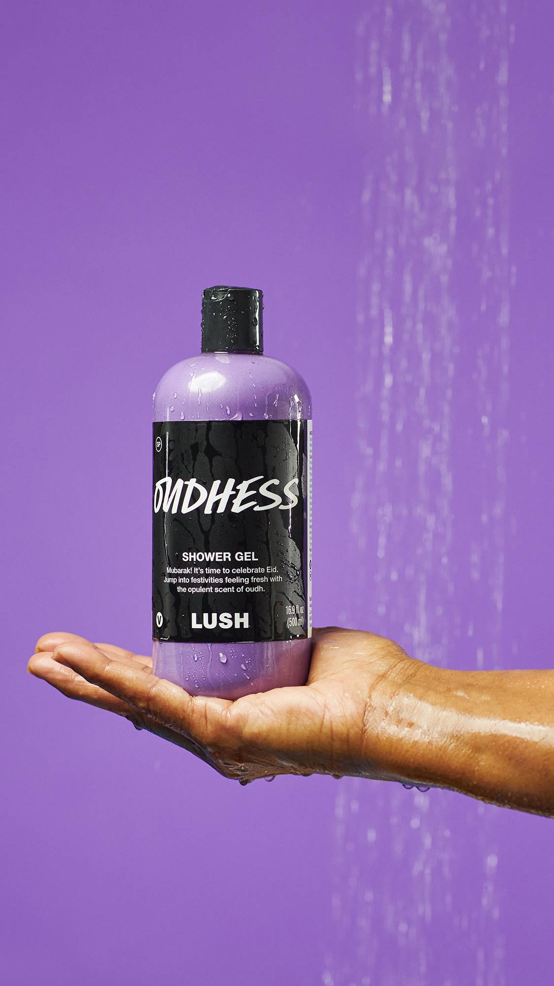 A close-up of the model's hand under running shower water as they hold an upright bottle of the Oudhess shower gel in their flat palm on a lilac background.