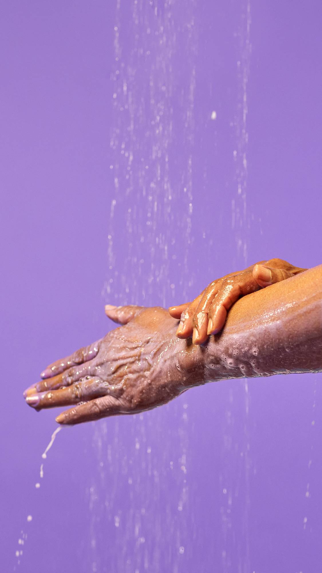 A close-up of the model's hands under running shower water as they are lathered up in thick soaps suds of Oudhess shower gel, on a lilac purple background. 