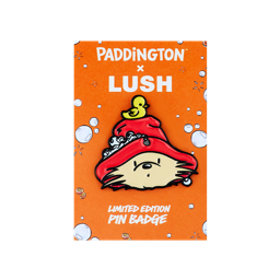 An enamel pin badge from the Paddington x Lush collection. The badge features Paddington Bear’s head wearing a red hat with a small yellow duck on top. It is displayed on an orange backing card with white bubbles and orange slice illustrations.