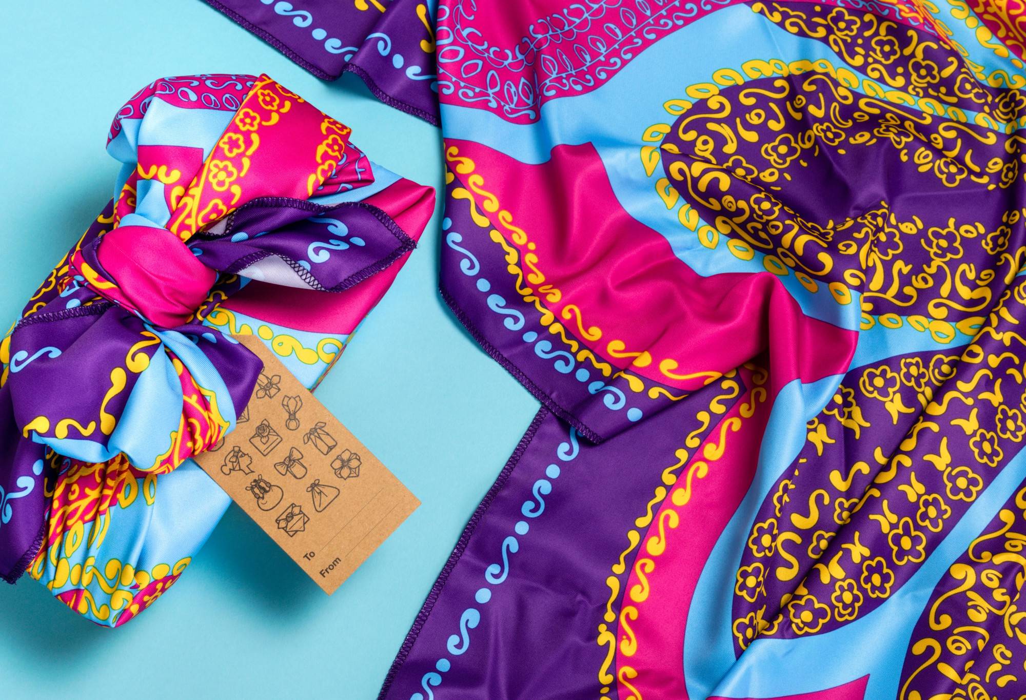 The knot wrap is wrapped around a rectangular box with a tag, as well as laid out flat, on a bright blue background.