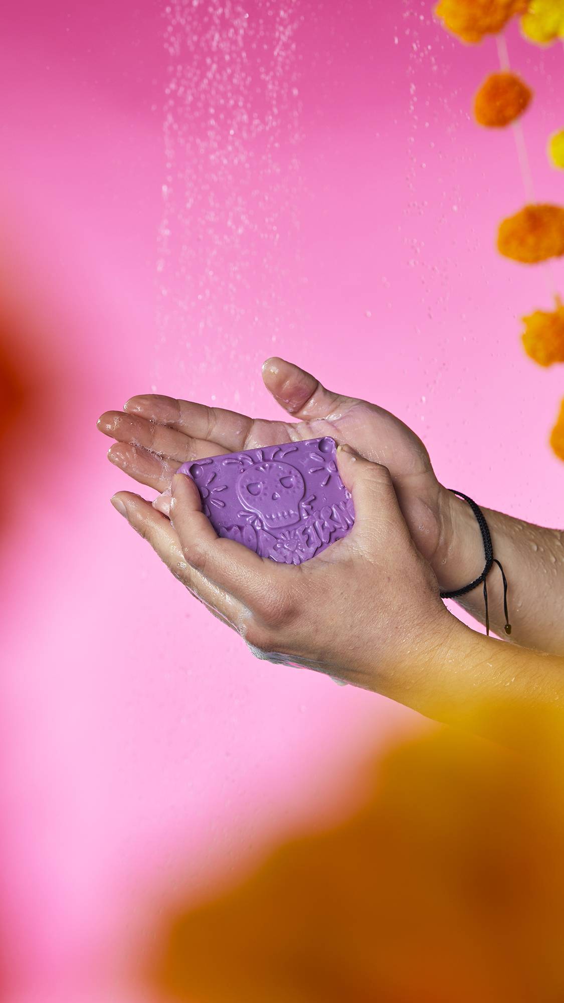 The image shows a pair of hands holding the purple Papel Picado Morado soap with a sugar skull shape embossed on top. Bubbly lather and water droplets gather in the cupped palms, against a vibrant pink background.