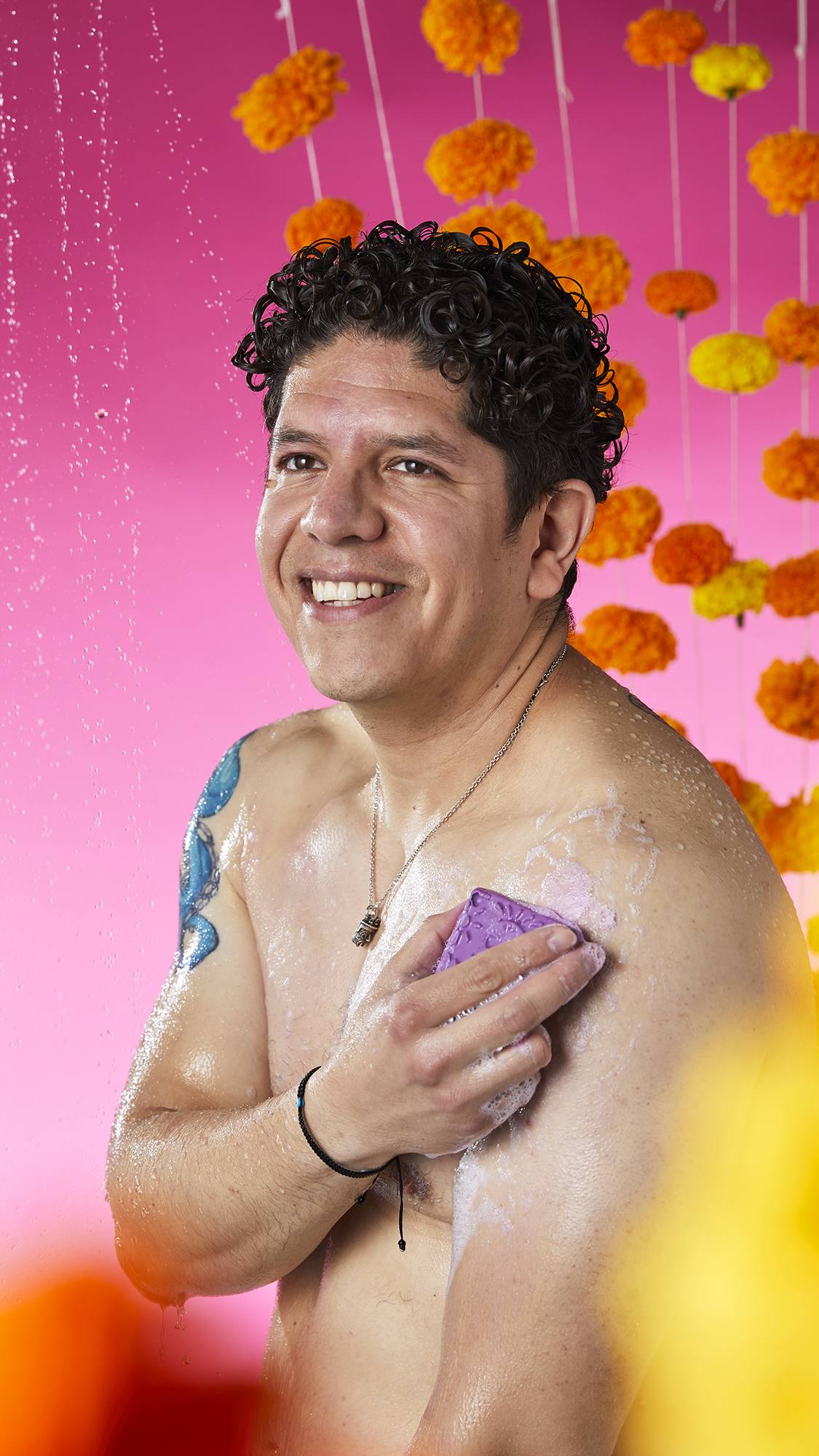 The image shows a person with a serene expression, rubbing a purple Papel Picado Morado Soap onto their shoulder. A bubbly lather is visible on the skin, against a vibrant pink background with flower garlands.