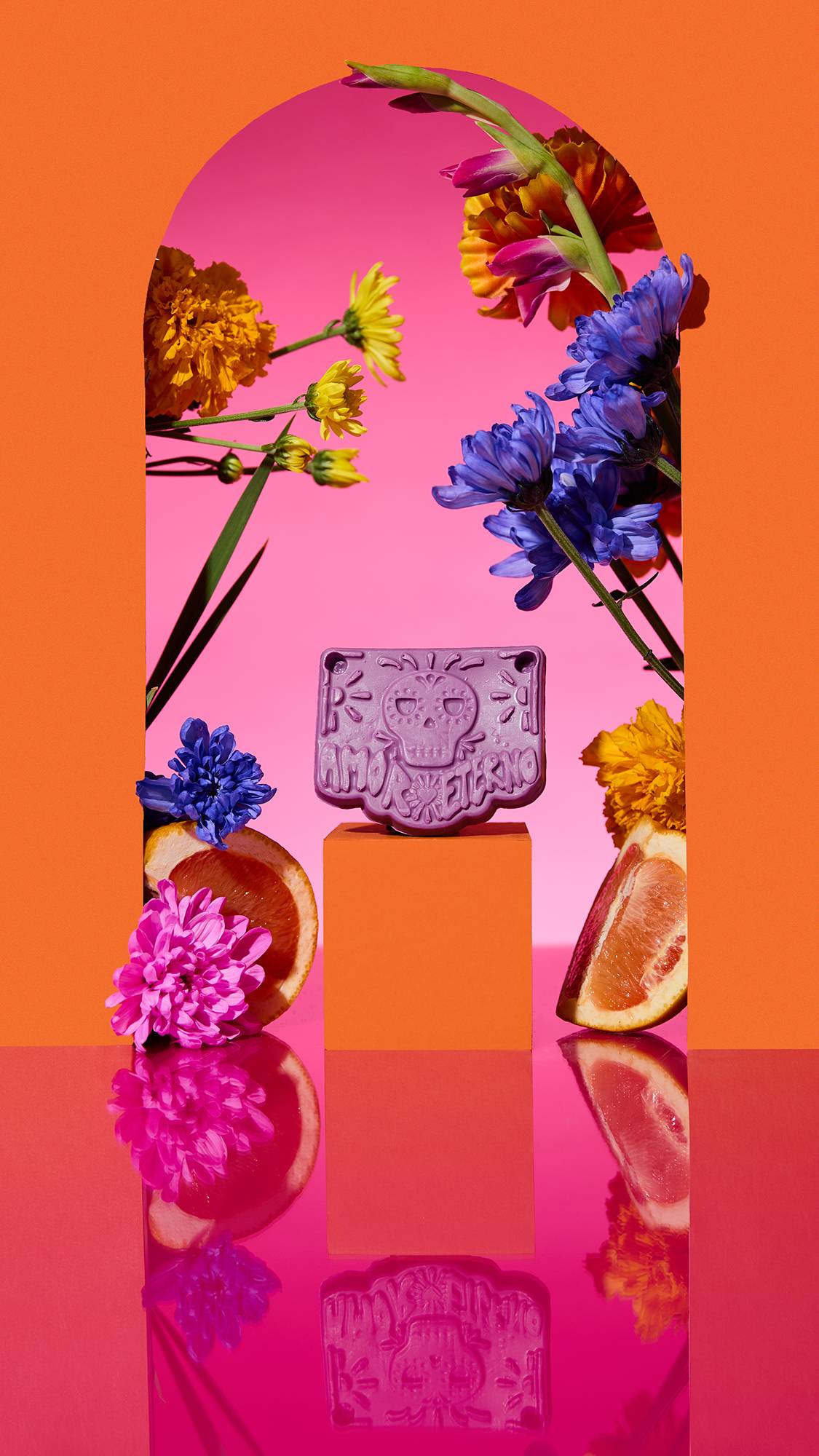 The image shows the purple Papel Picado Morado Soap on a plinth. A colourful arch frames the scene, with sliced citrus, flowers, and a cinnamon stick surrounding the product on a reflective pink surface.