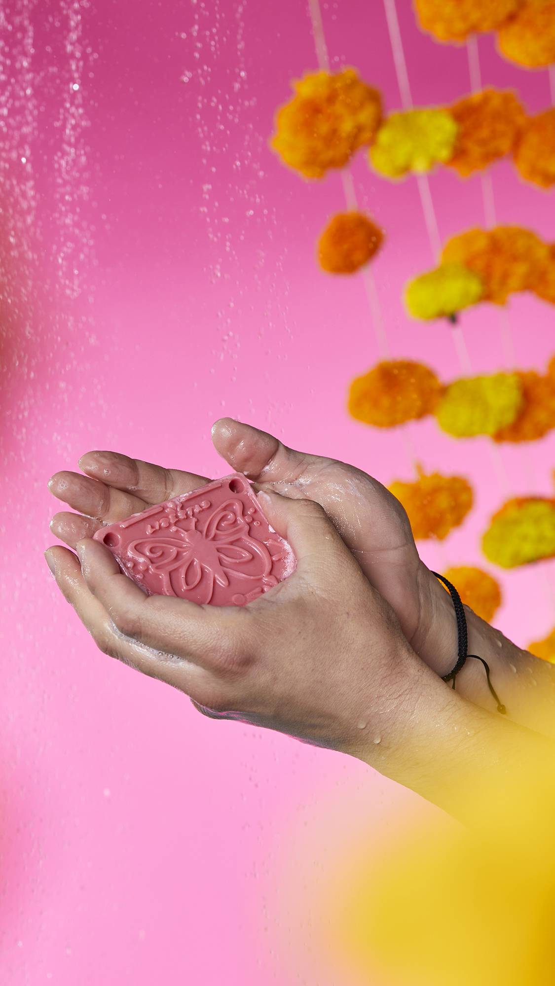 The image shows a pair of hands holding the pink Papel Picado Rosa soap with a butterfly shape embossed on top. Bubbly lather and water droplets gather in the cupped palms, against a vibrant pink background.