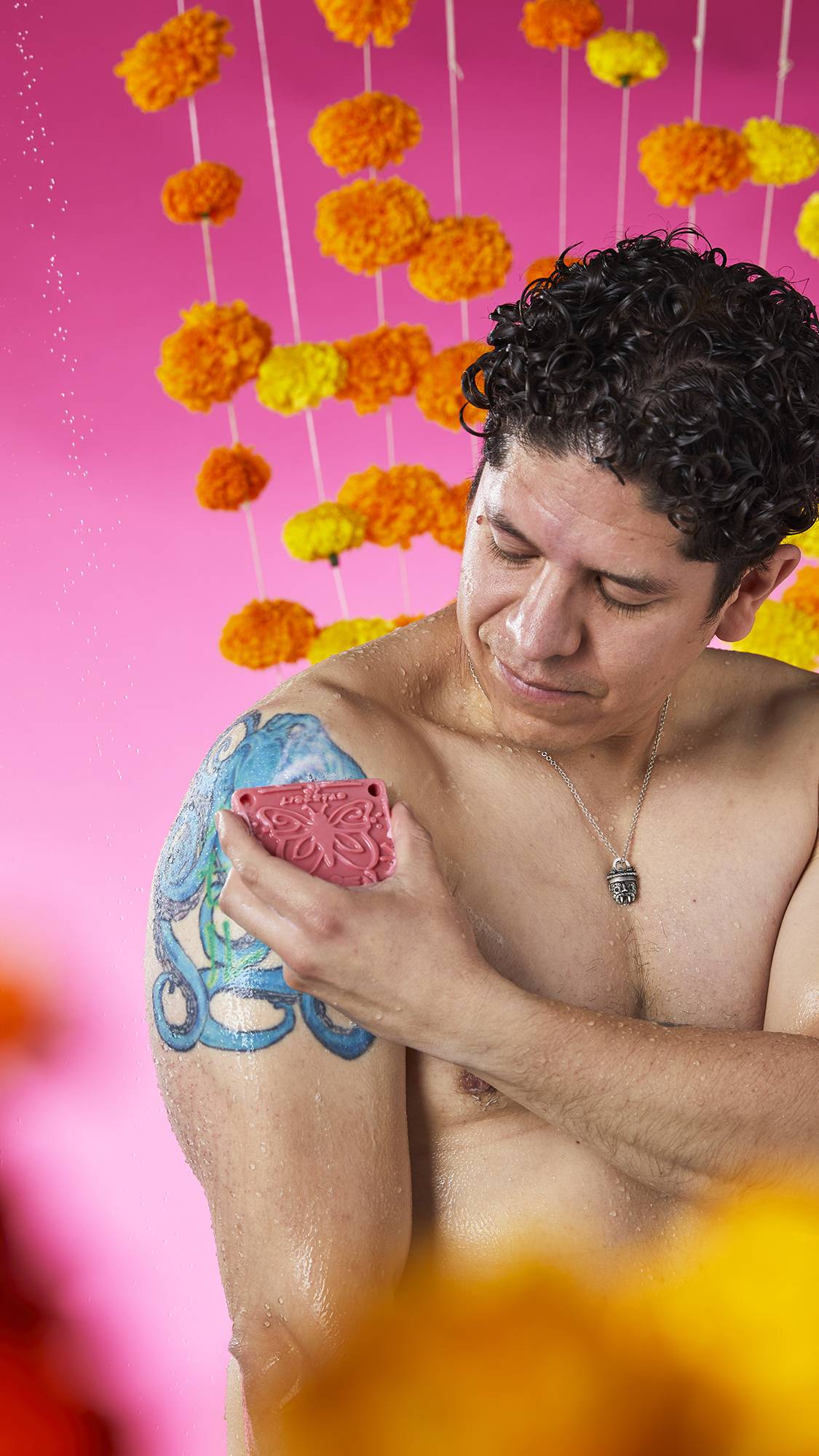 The image shows a person with a serene expression, rubbing a pink Papel Picado Rosa Soap onto their tattooed shoulder. A bubbly lather is visible on the skin, against a vibrant pink background with flower garlands.