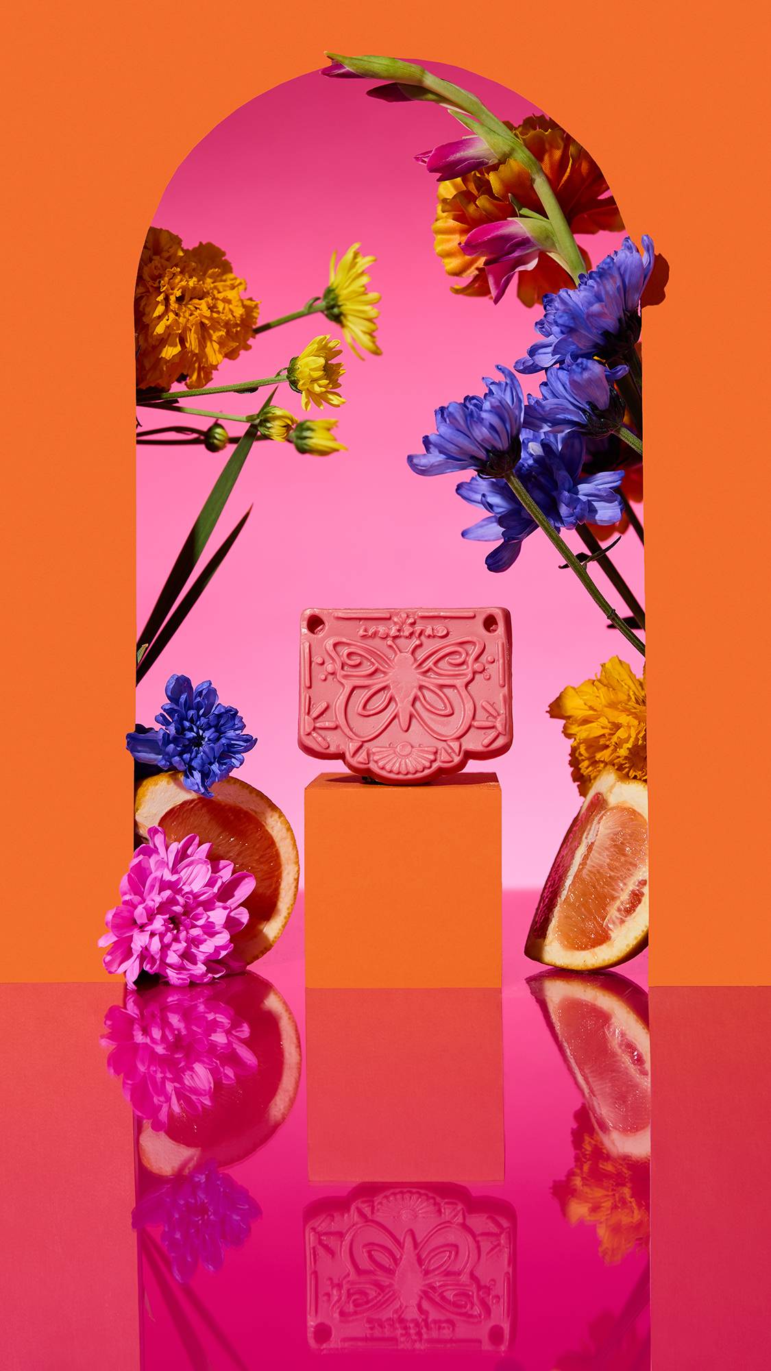The image shows the pink Papel Picado Rosa Soap on a plinth. A colourful arch frames the scene, with sliced citrus, flowers, and a cinnamon stick surrounding the product on a reflective pink surface.