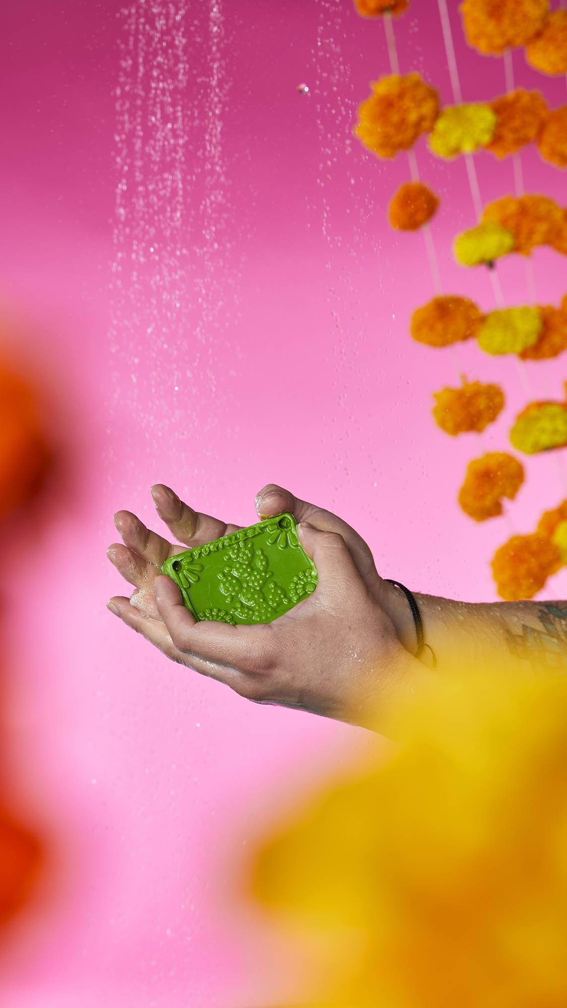 The image shows a pair of hands holding the green Papel Picado Verde soap with a cactus shape embossed on top. Bubbly lather and water droplets gather in the cupped palms, against a vibrant pink background.