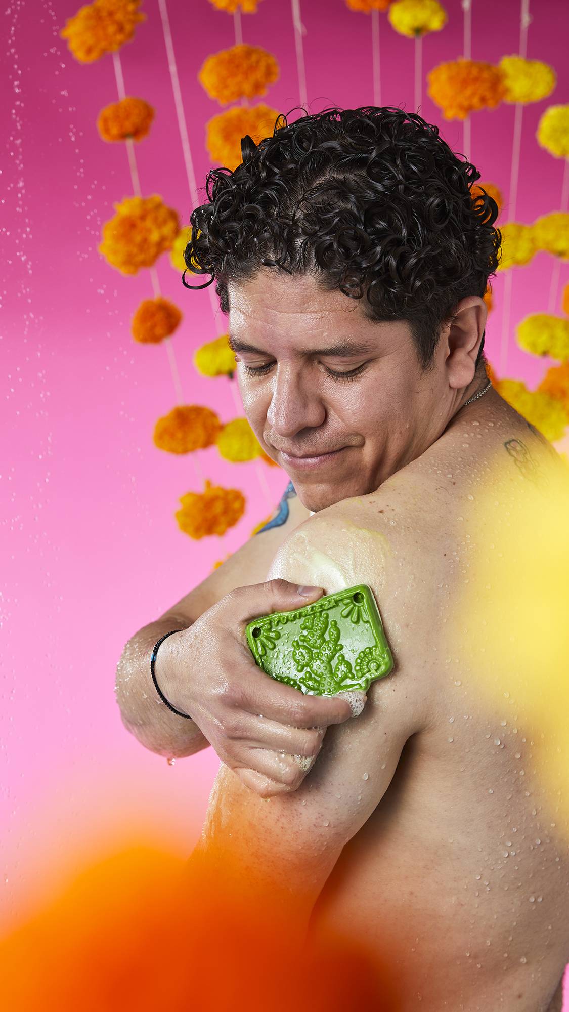 The image shows a person with a serene expression, rubbing a green Papel Picado Verde Soap onto their shoulder. A bubbly lather is visible on the skin, against a vibrant pink background with flower garlands.