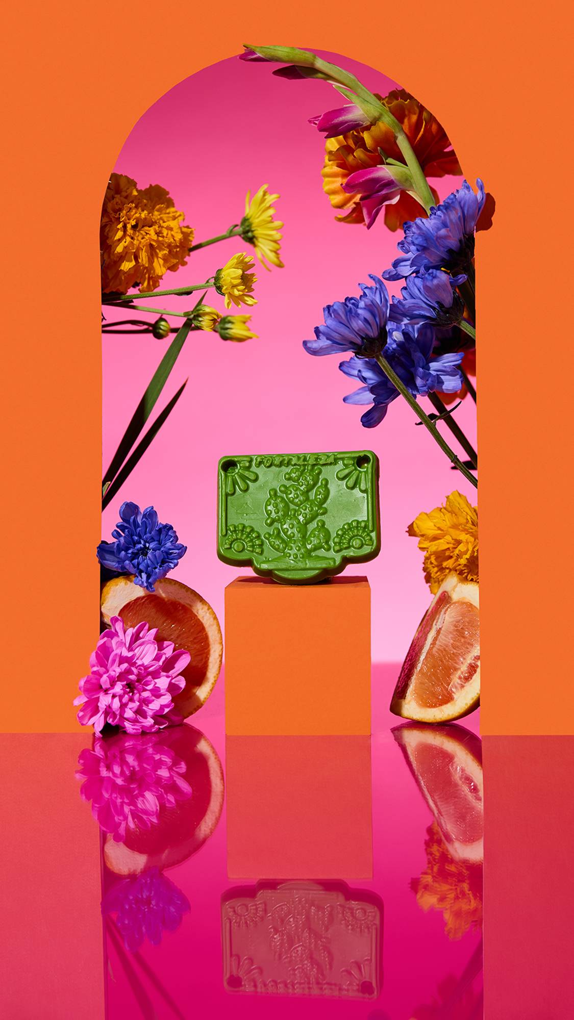 The image shows the bright green Papel Picado Verde Soap on a plinth. A colourful arch frames the scene, with sliced citrus, flowers, and a cinnamon stick surrounding the product on a reflective pink surface.