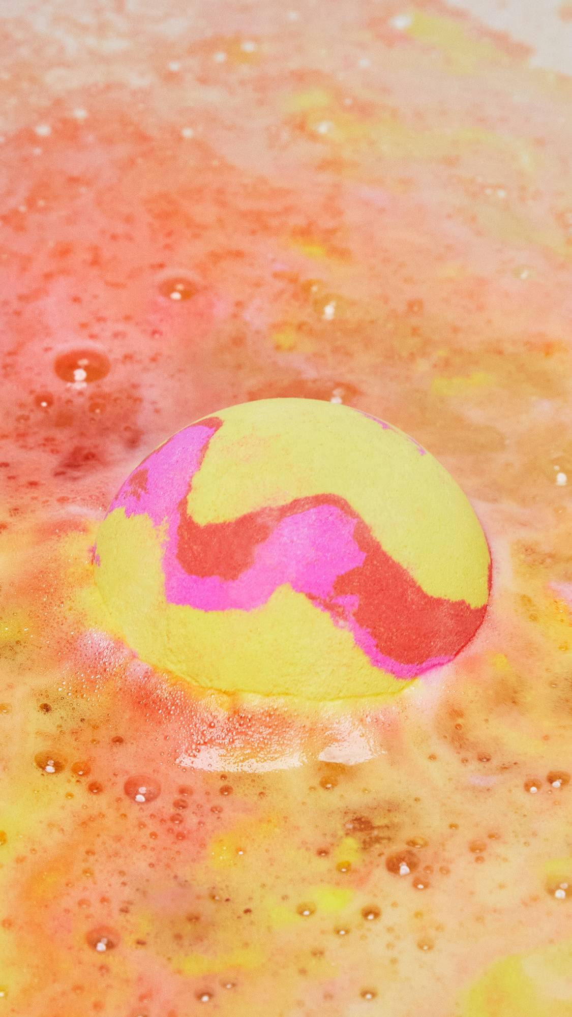 The Passion bath bomb is sitting on top of the water slowly dissolving creating swirling ripples of yellow, pink and orange. 