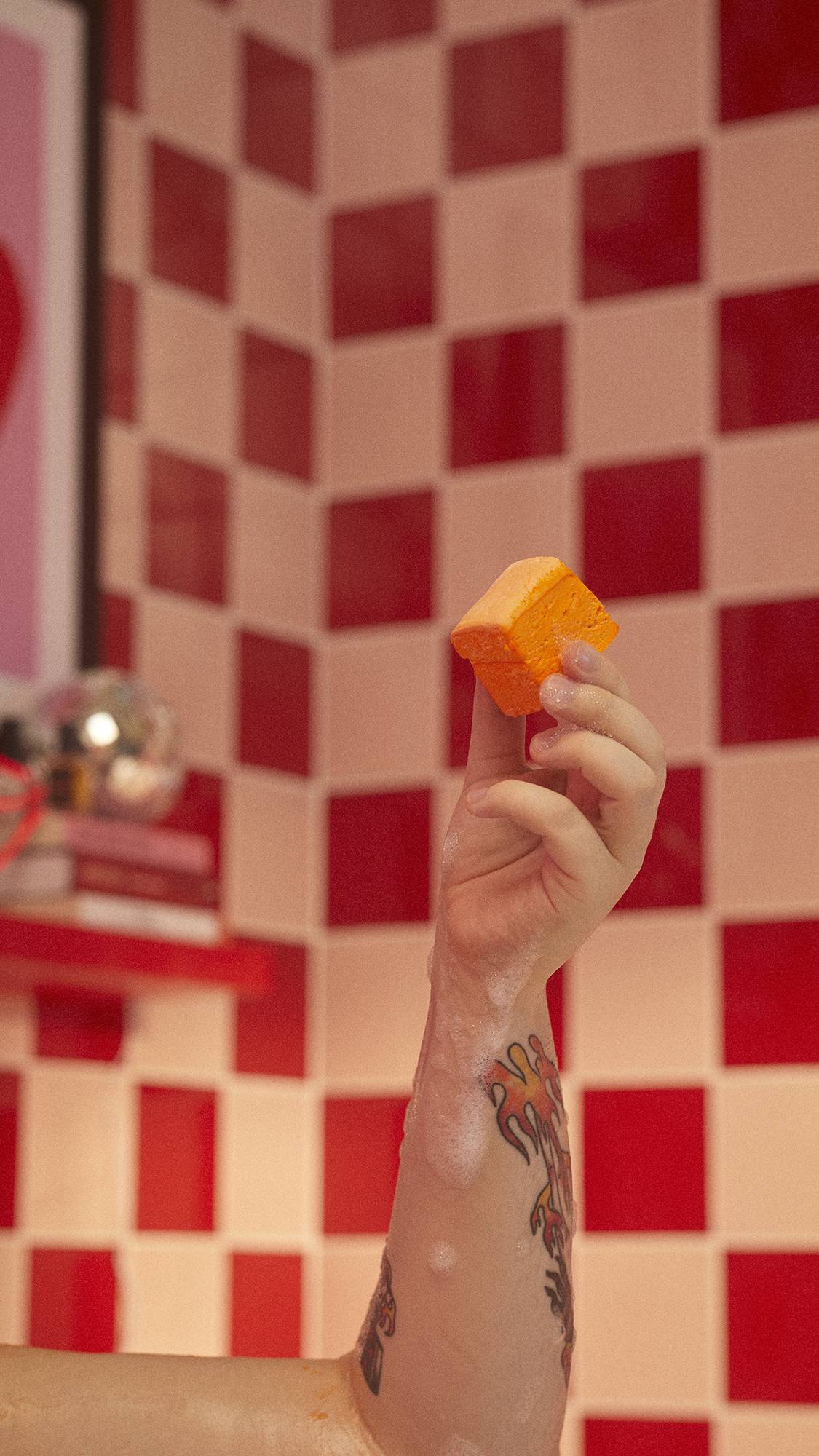 A close-up image of the model's hand holding the solid, cube Passion Fruit Delight body was in the air, in a tiled bathroom.