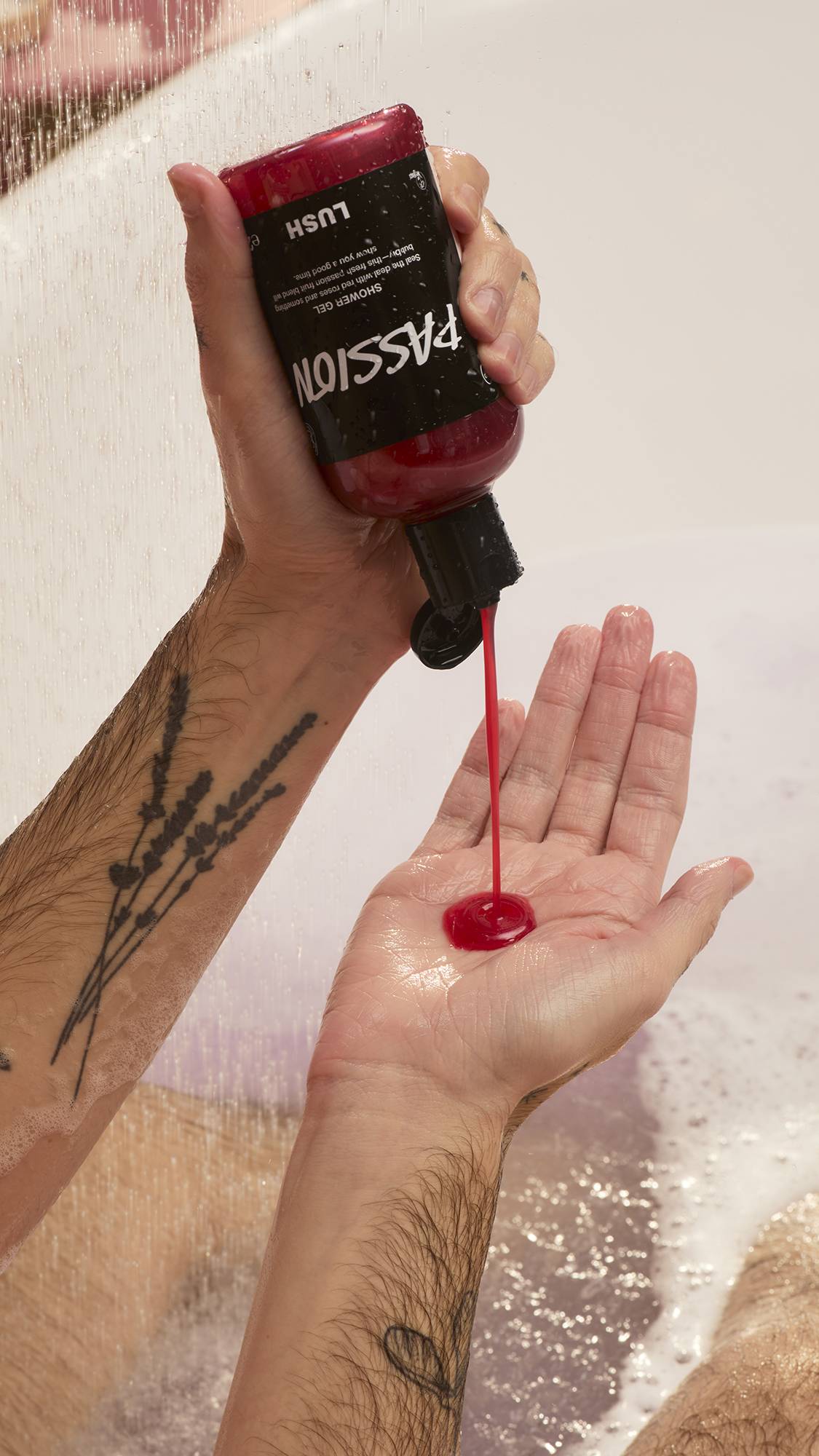 A model with tattooed arms squeezes a bottle of the deep red Lush "Passion" shower gel into their hand while in a bathtub. The vibrant gel streams from the bottle as water falls in the background, creating a soapy lather in the tub.