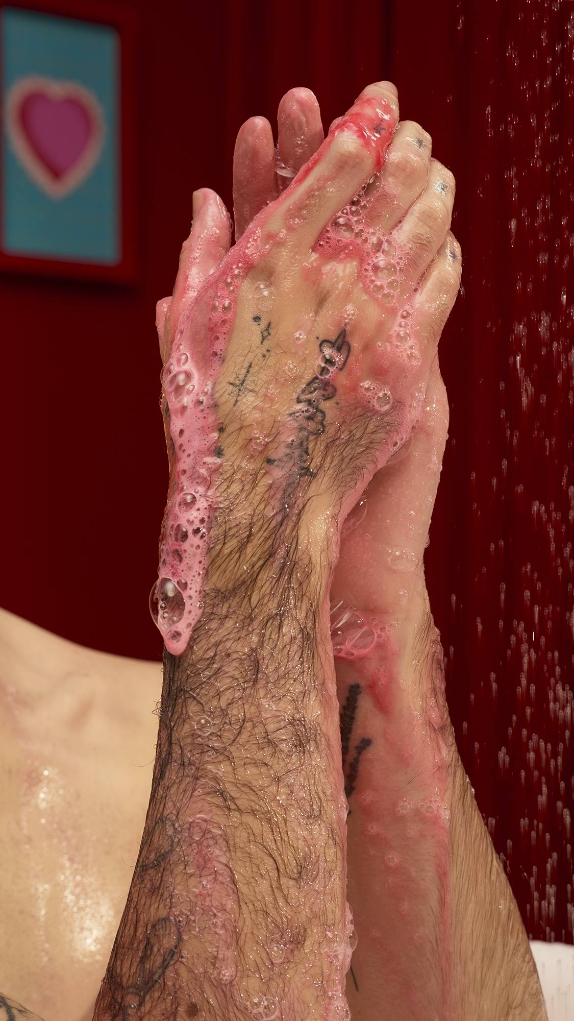 A person with tattooed arms lathers their hands under a shower stream. Bright pink soap bubbles from Lush "Passion" shower gel cover their wet skin and several black ink tattoos on their forearms against a deep red background.