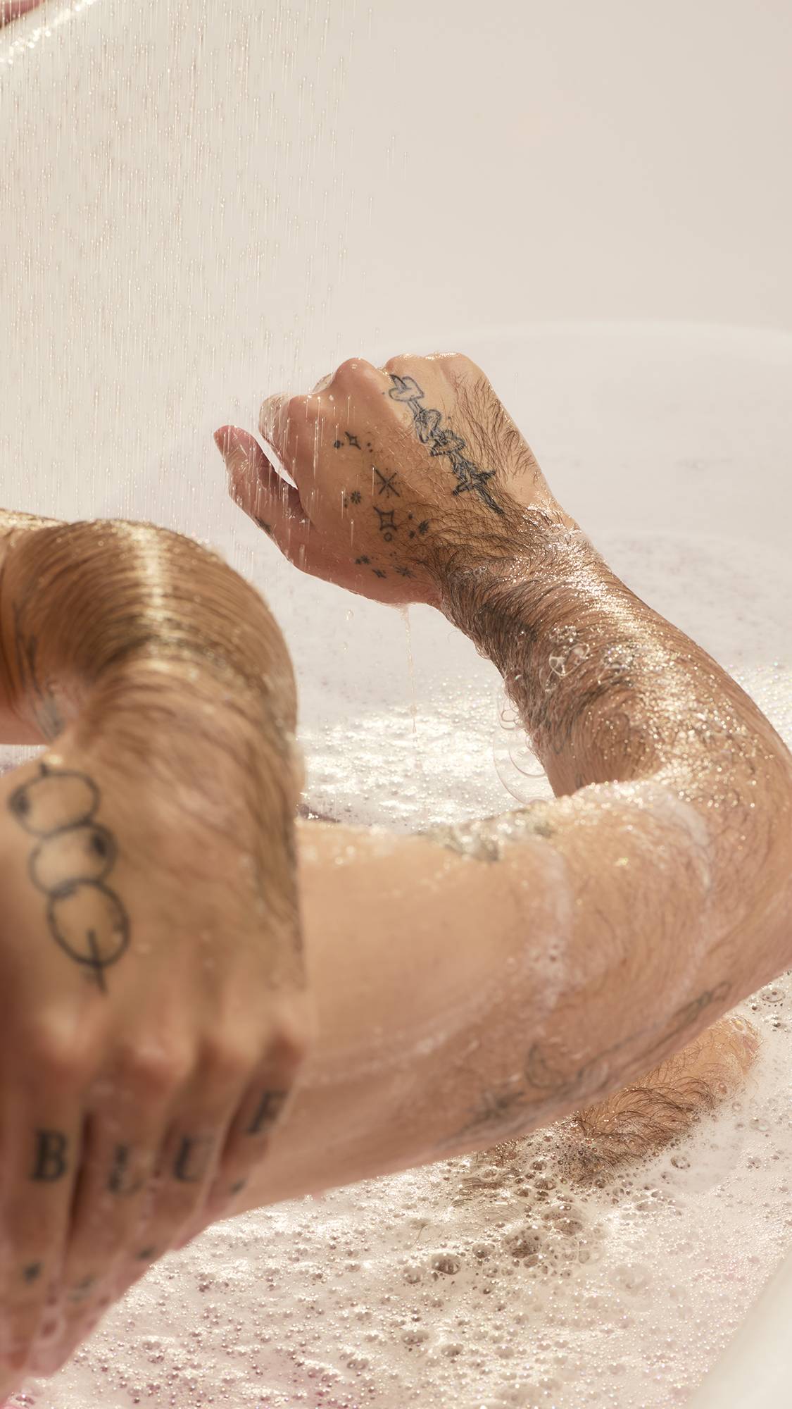 A person with tattooed arms washes their hands and forearms in a white bathtub. Their skin is covered in white soap bubbles under a falling stream of water, which also fills the tub with foamy, slightly pink bathwater from the Passion shower gel.