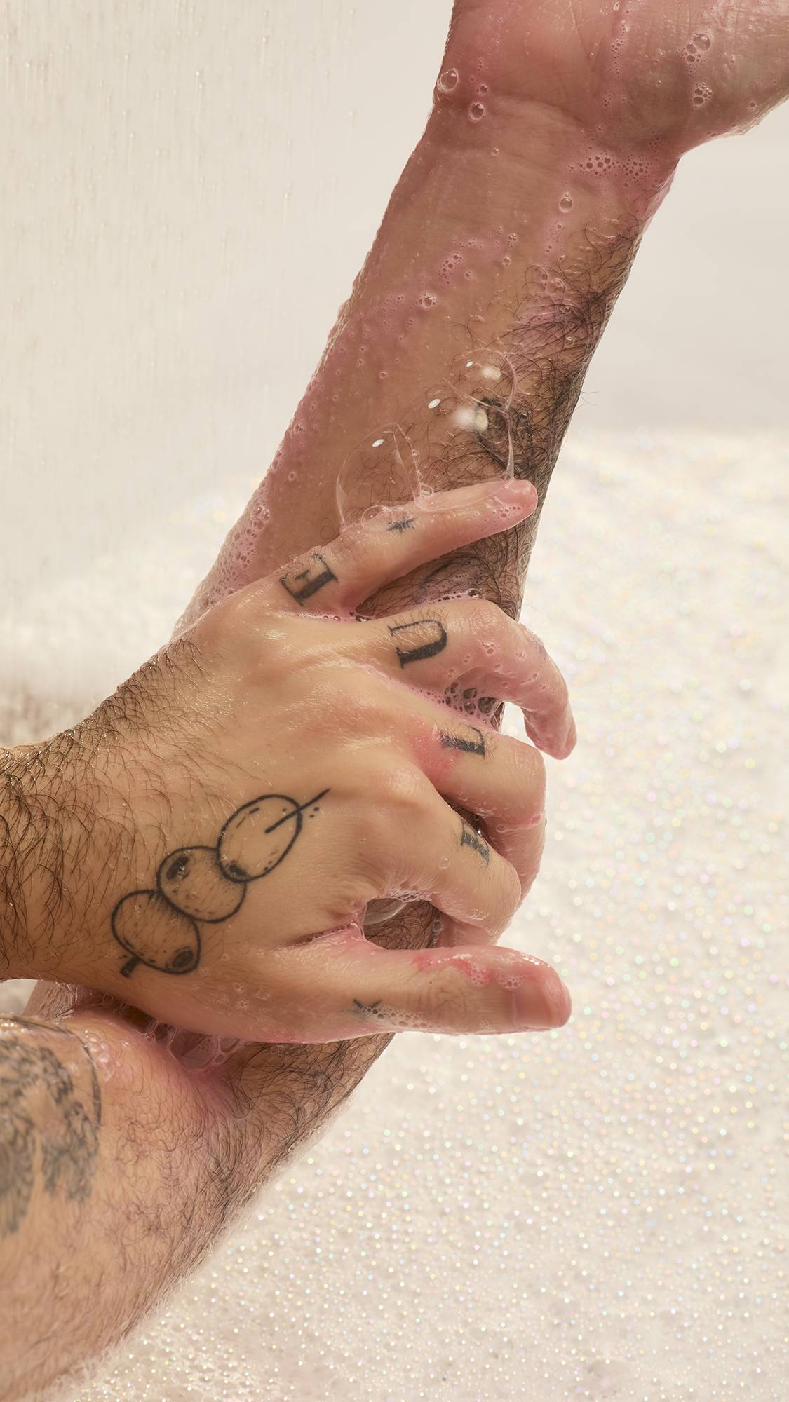 A person with tattooed arms lathers their hand and forearm in a bathtub. Pink soap suds from Lush "Passion" shower gel cover their wet skin and several black ink tattoos under a stream of water against a foamy background.