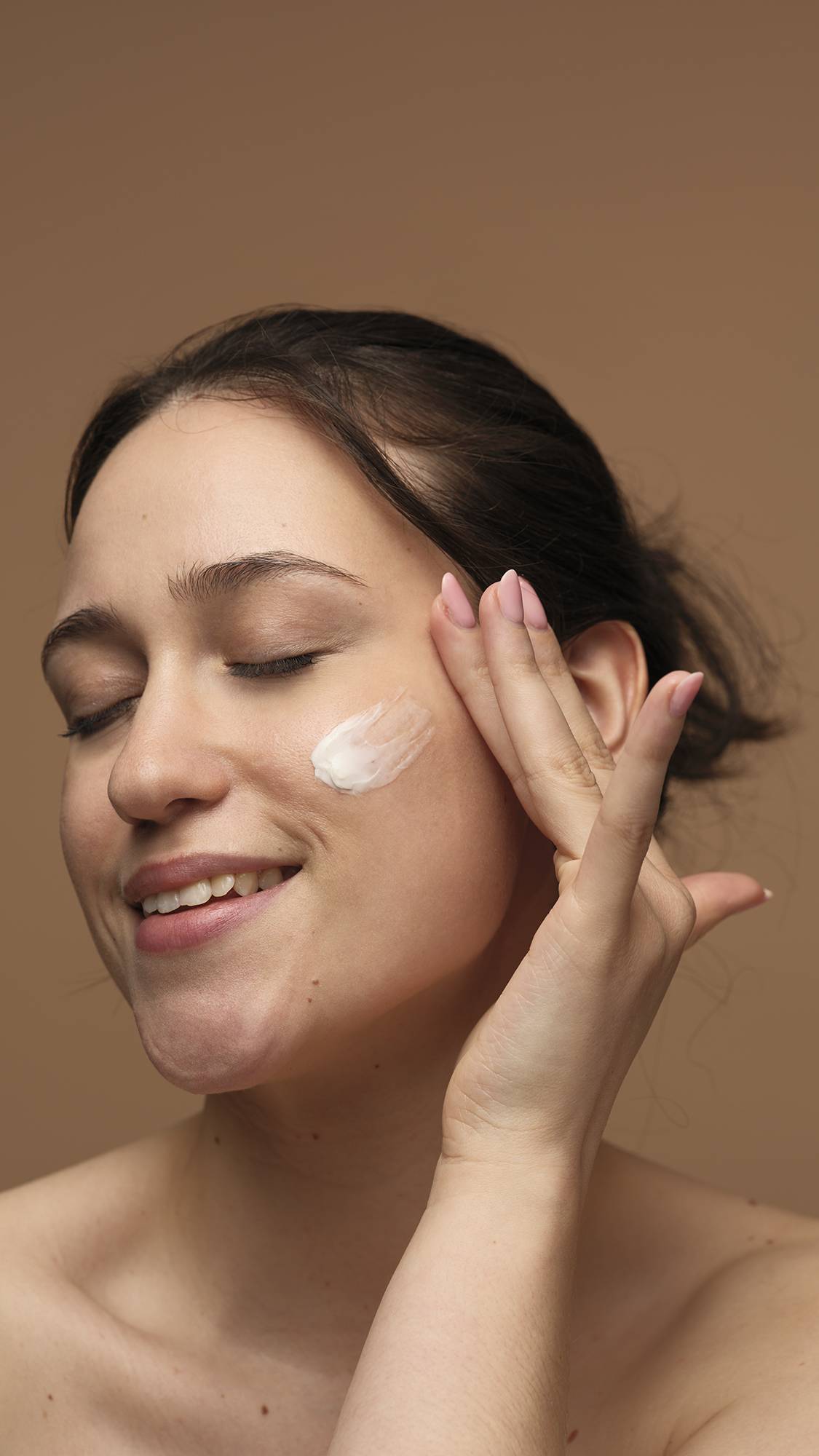 The image shows the model's face as their eyes are closed while they gently sweep the Peace moisturiser over their cheek. They are on a warm, earthy-brown background.