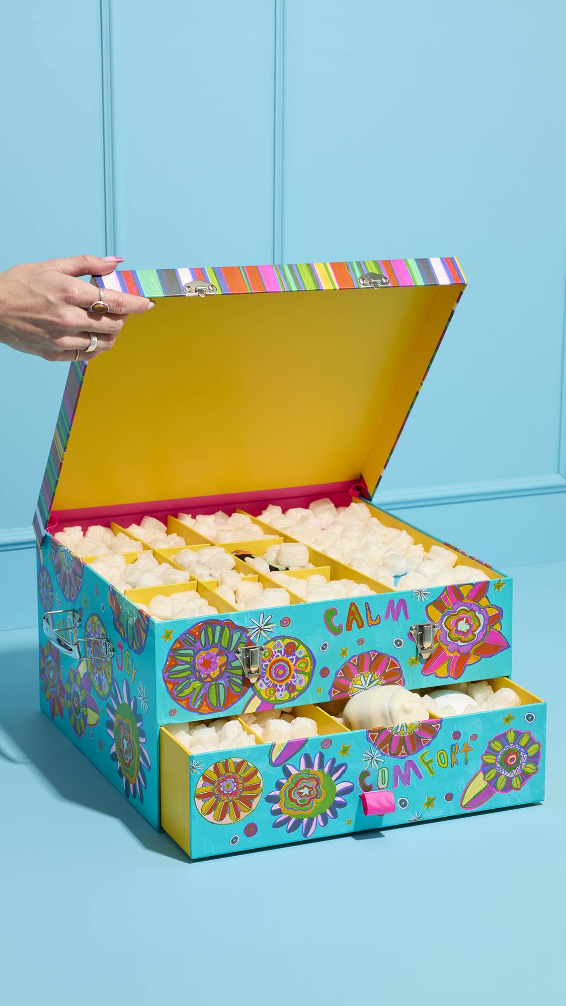A hand opens the Peace and Love gift trunk, showing a yellow lid and compartments filled with white eco flow. The bottom 'COMFORT' drawer is pulled out, revealing more packing material and a scubee