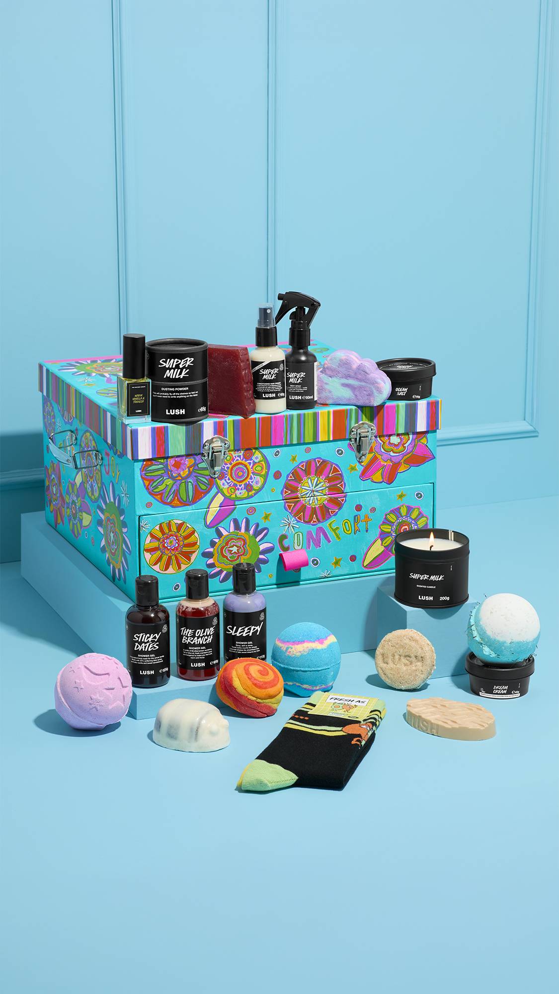 The Peace and Love gift features a large blue floral trunk with a "COMFORT" drawer. It is surrounded by Lush products: shower gels, bath bombs, a Super Milk candle, and socks, all on a light blue background.