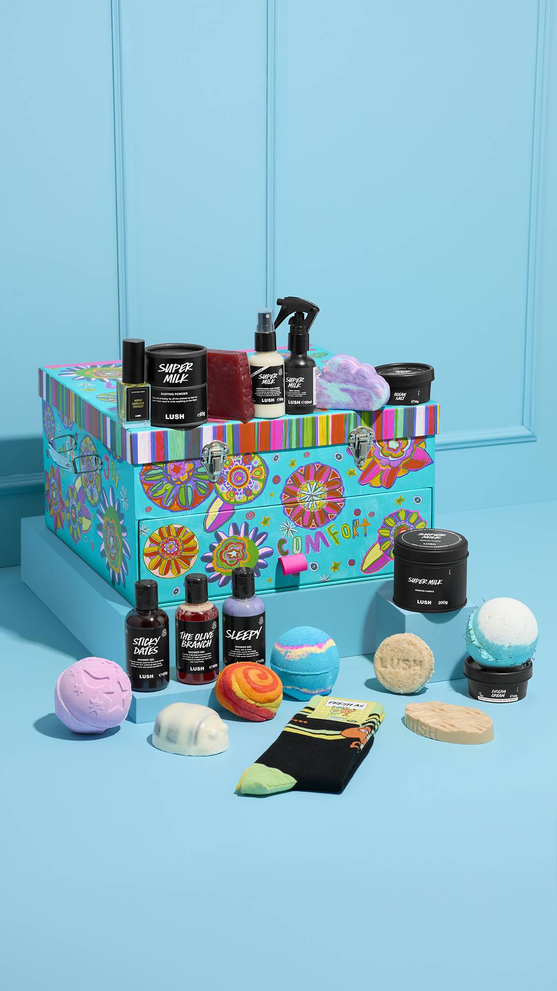 The Peace and Love gift is a large, colorful floral trunk with a 'COMFORT' drawer. It is surrounded by Lush products like bath bombs, shower gels, and socks on a light blue background.