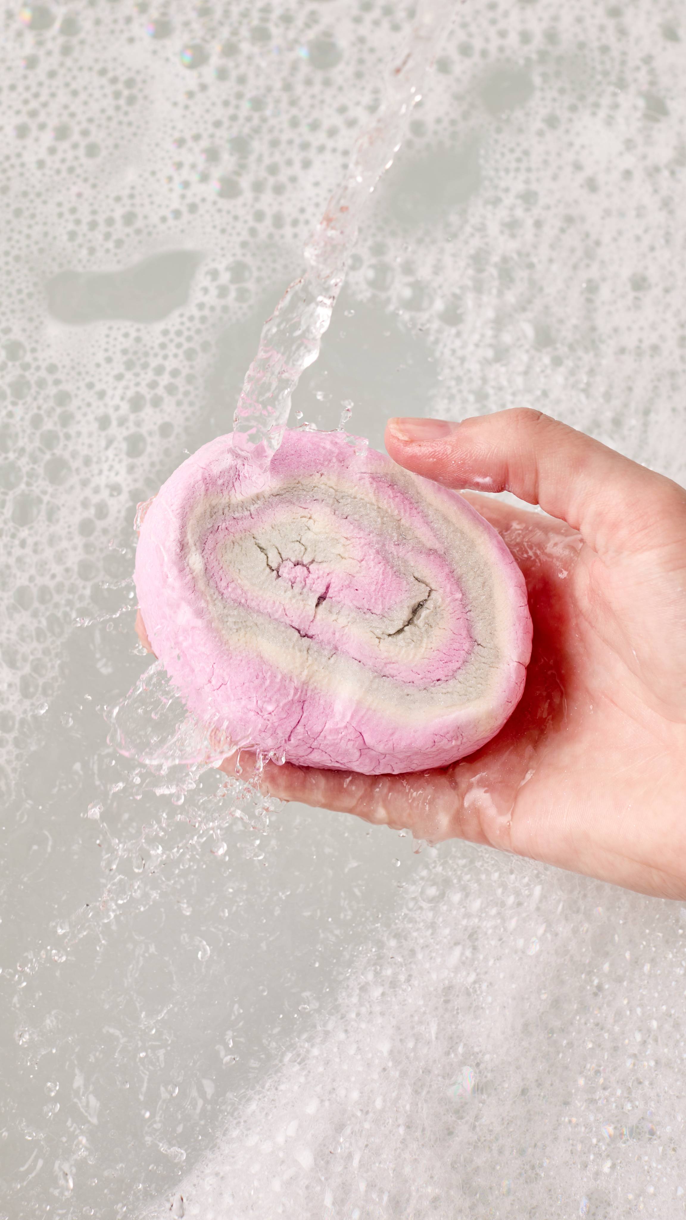 The image shows a light-toned hand holding a pink and white 'Phantom Foam' bubble bar with a spiral pattern. Water pours onto the bar, generating abundant white bubbles and thick foam across the bath.