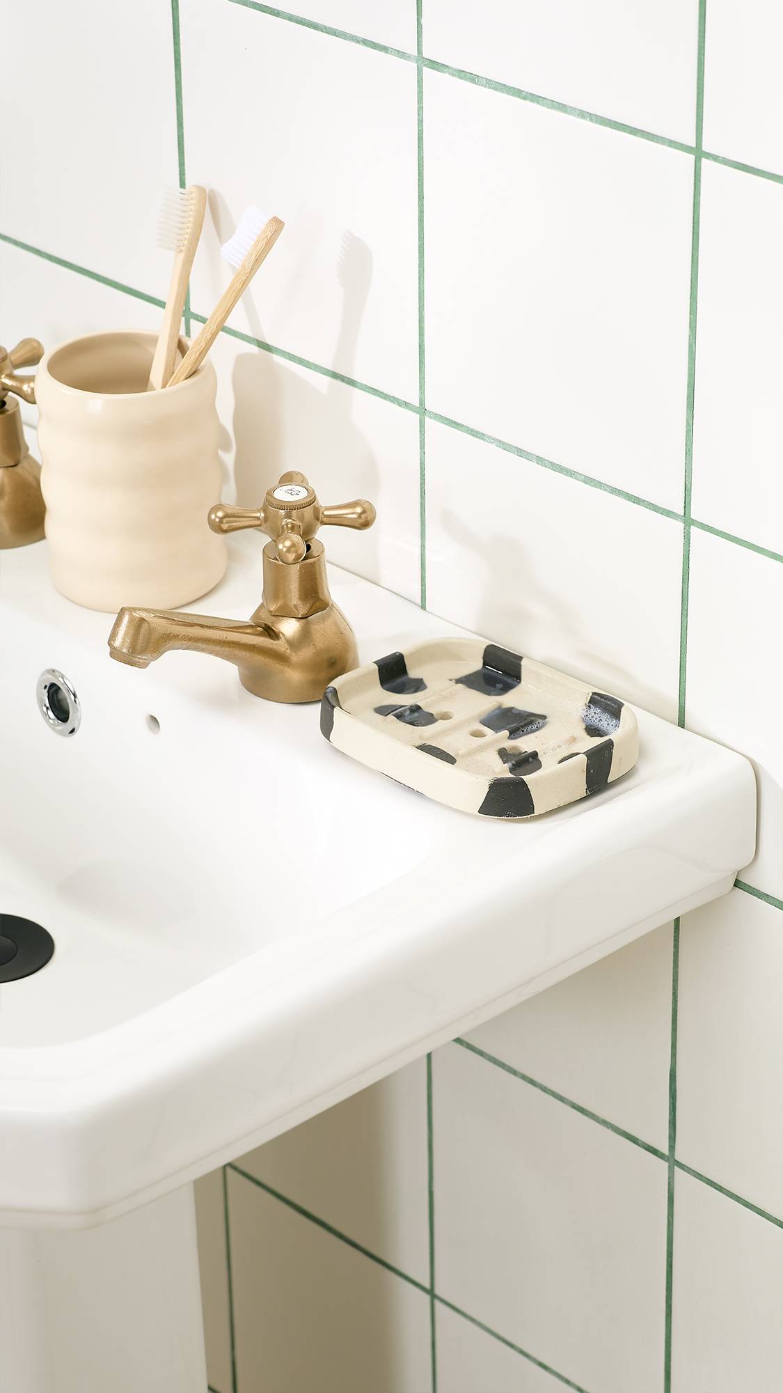 A cream and black cow-print ceramic Lush "Phoenix" soap dish sits beside a brass tap on a white pedestal sink, next to a ceramic holder with wooden toothbrushes set against a white tiled wall.   