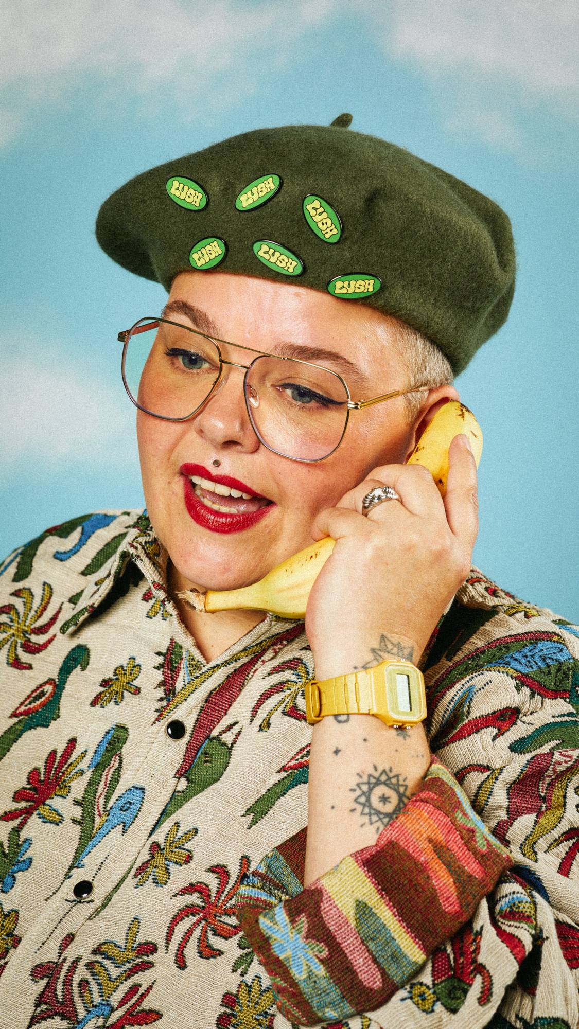 Model wears a bold shirt as they hold a banana to their face like a phone. They're wearing a green beret with multiple badges.