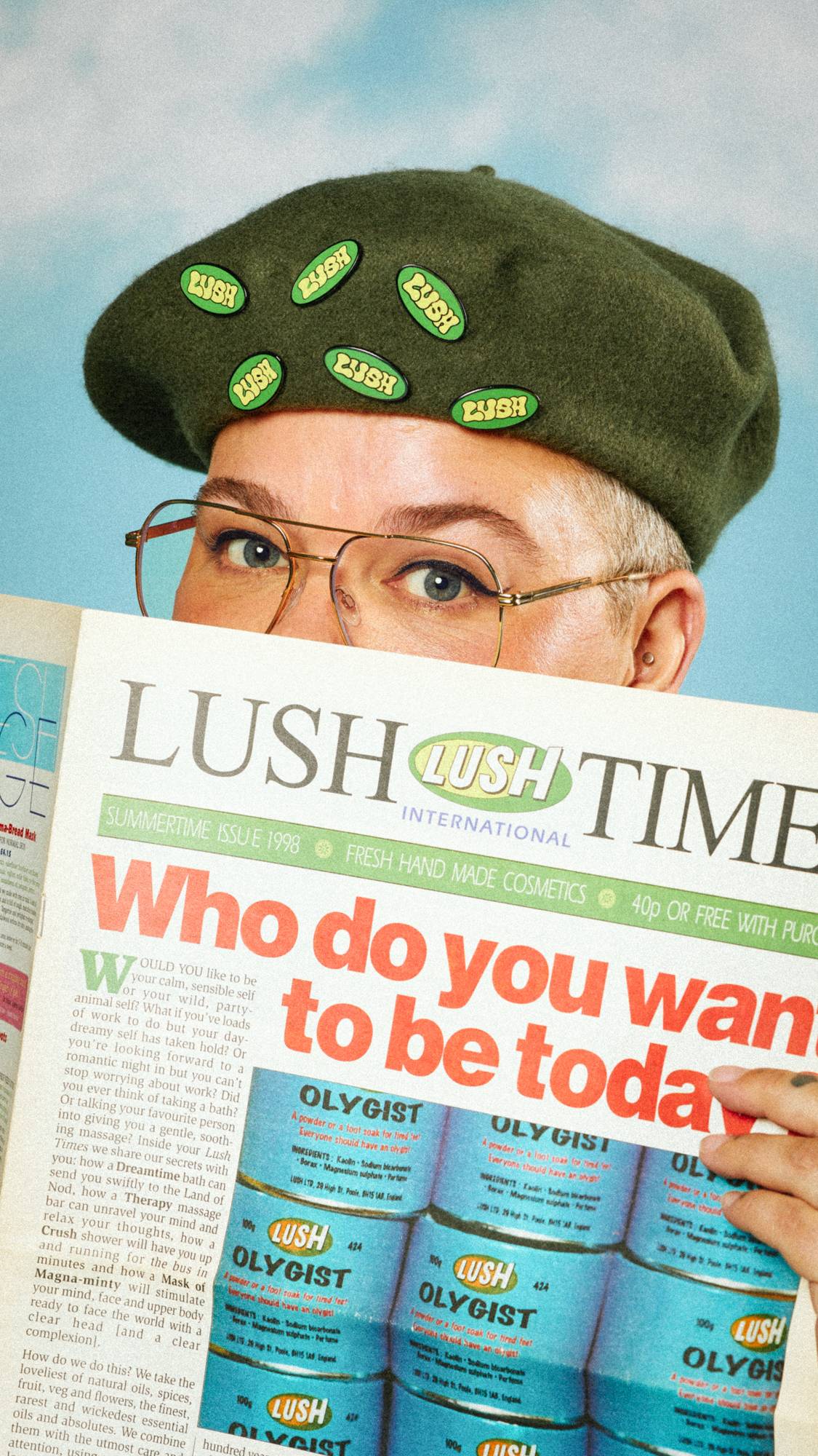 Model is holding The Lush Times magazine up as their glasses peer over. They're wearing a green beret with multiple badges on.