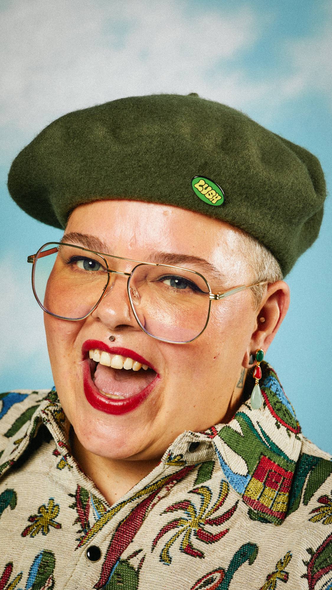 Model is wearing a bold shirt with glasses and a green beret hat with a Retro Bubble Lush pin badge attached to the front. 