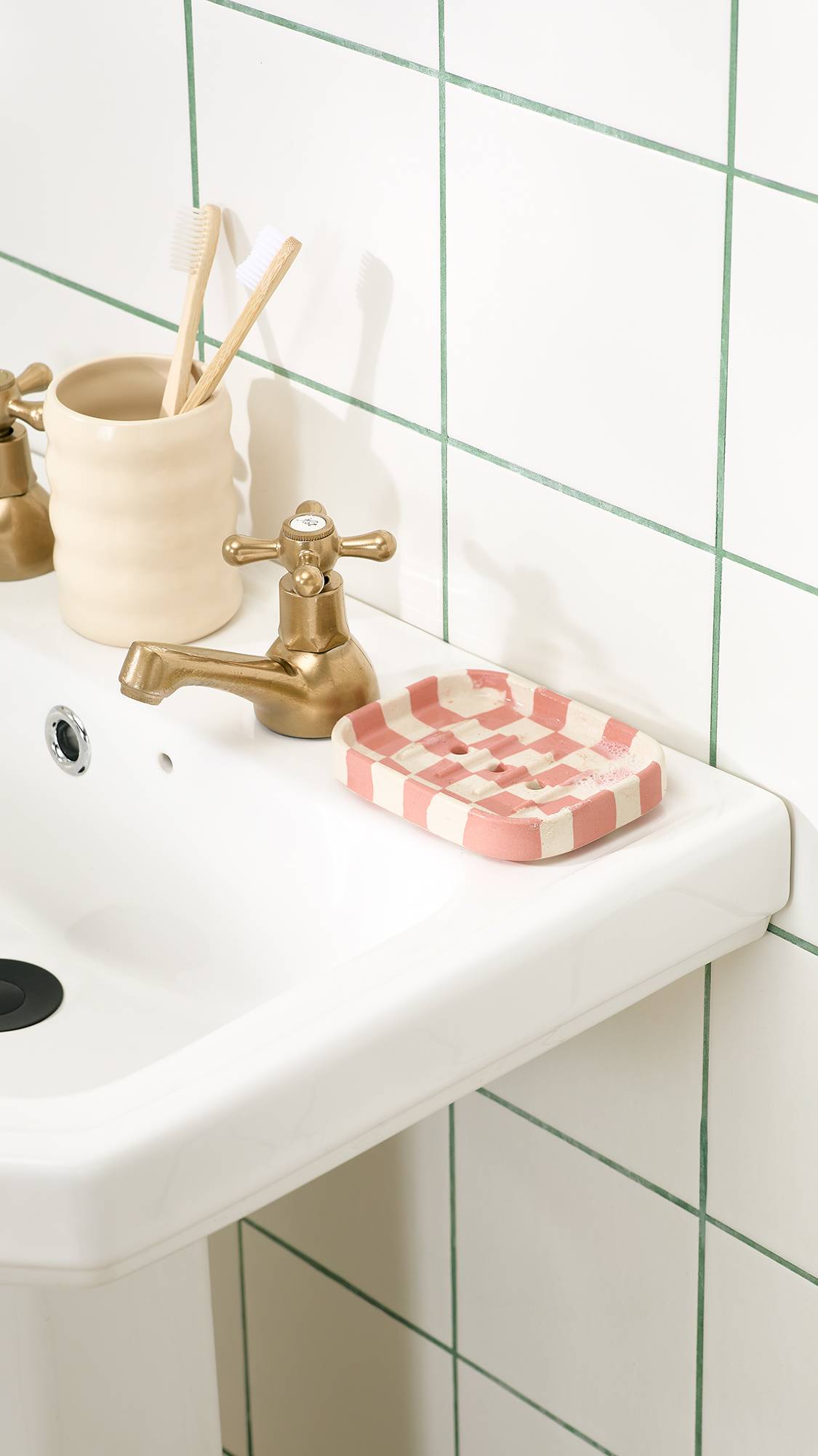 A pink and cream chequered ceramic soap dish sits beside a brass tap on a white pedestal sink, next to a ceramic holder with wooden toothbrushes set against a white tiled wall.   