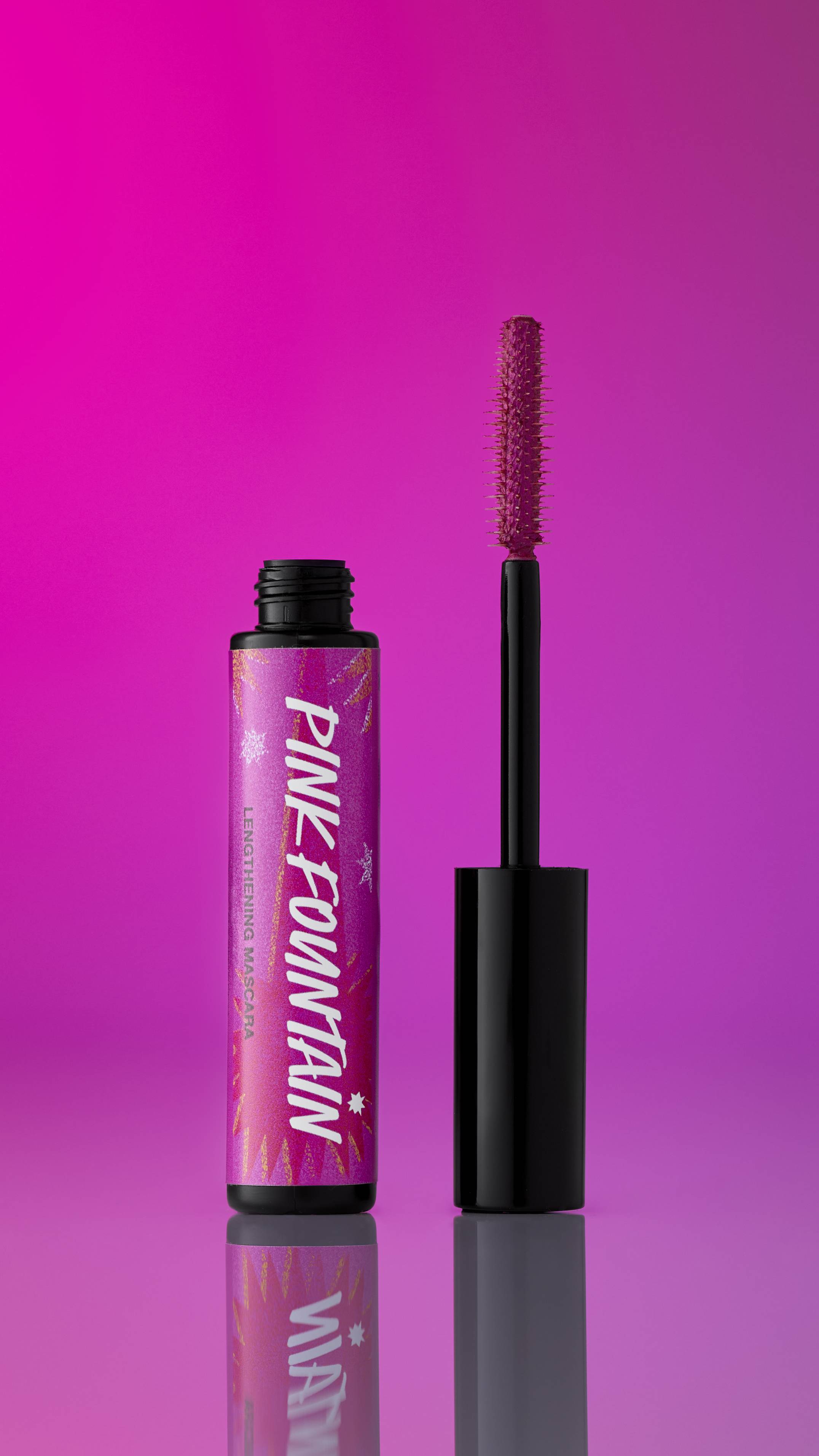 A black tube of Pink Fountain ~ Macara mascara stands with its cap unscrewed, showing the pink-bristled wand. The label is magenta with gold accents. It is set on a reflective surface against a gradient background of bright pink and magenta.