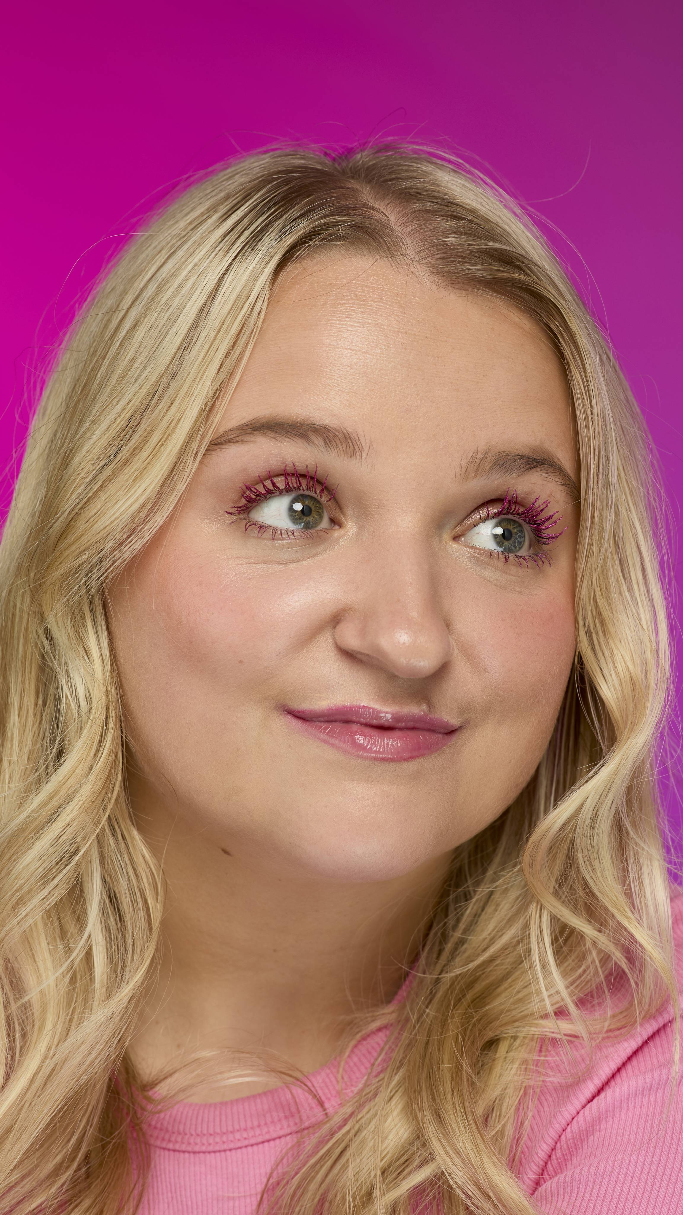 A person with blonde, wavy hair and a pink top looks up and to the side. They are wearing bright pink mascara on their eyelashes, consistent with Pink Fountain ~ Macara, set against a vivid magenta background.