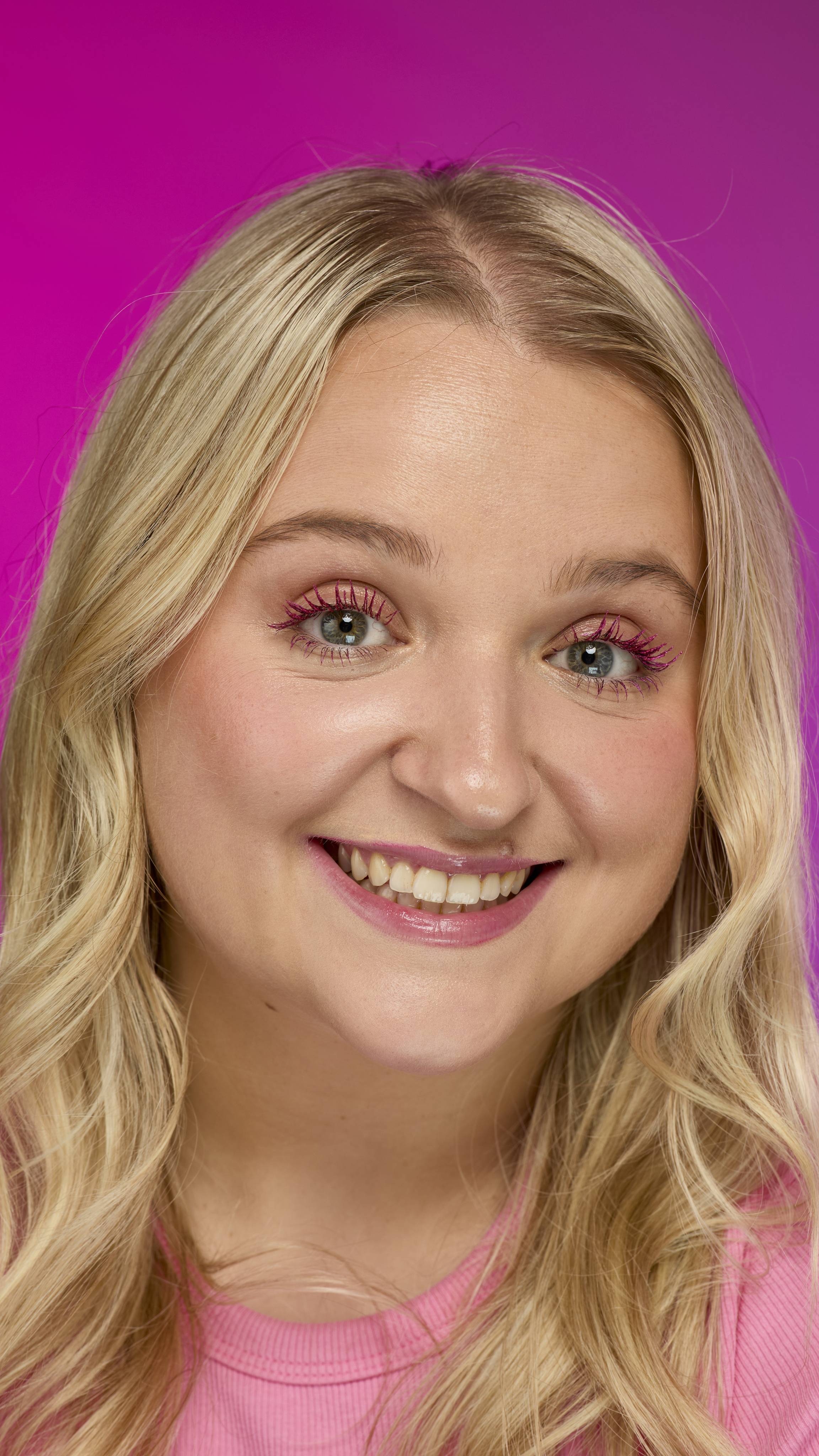 A person with blonde, wavy hair and a pink top smiles directly at the camera. They are wearing bright pink mascara on their eyelashes, consistent with Pink Fountain ~ Macara, set against a vivid magenta background.