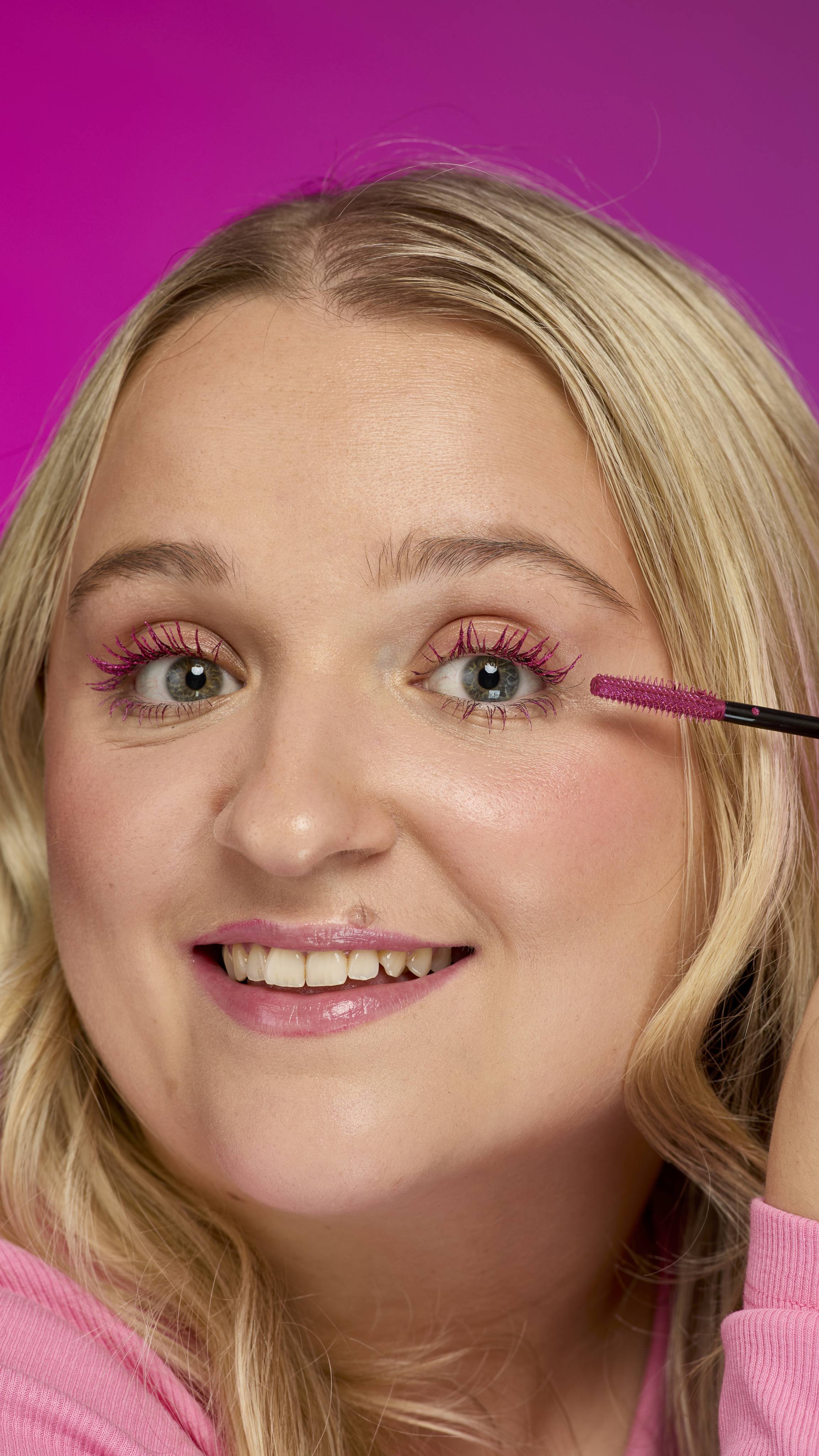 A person with blonde, wavy hair is applying Pink Fountain ~ Macara to their eyelashes with the pink-bristled mascara wand. They are wearing a pink top and smiling at the camera, against a vivid magenta background.