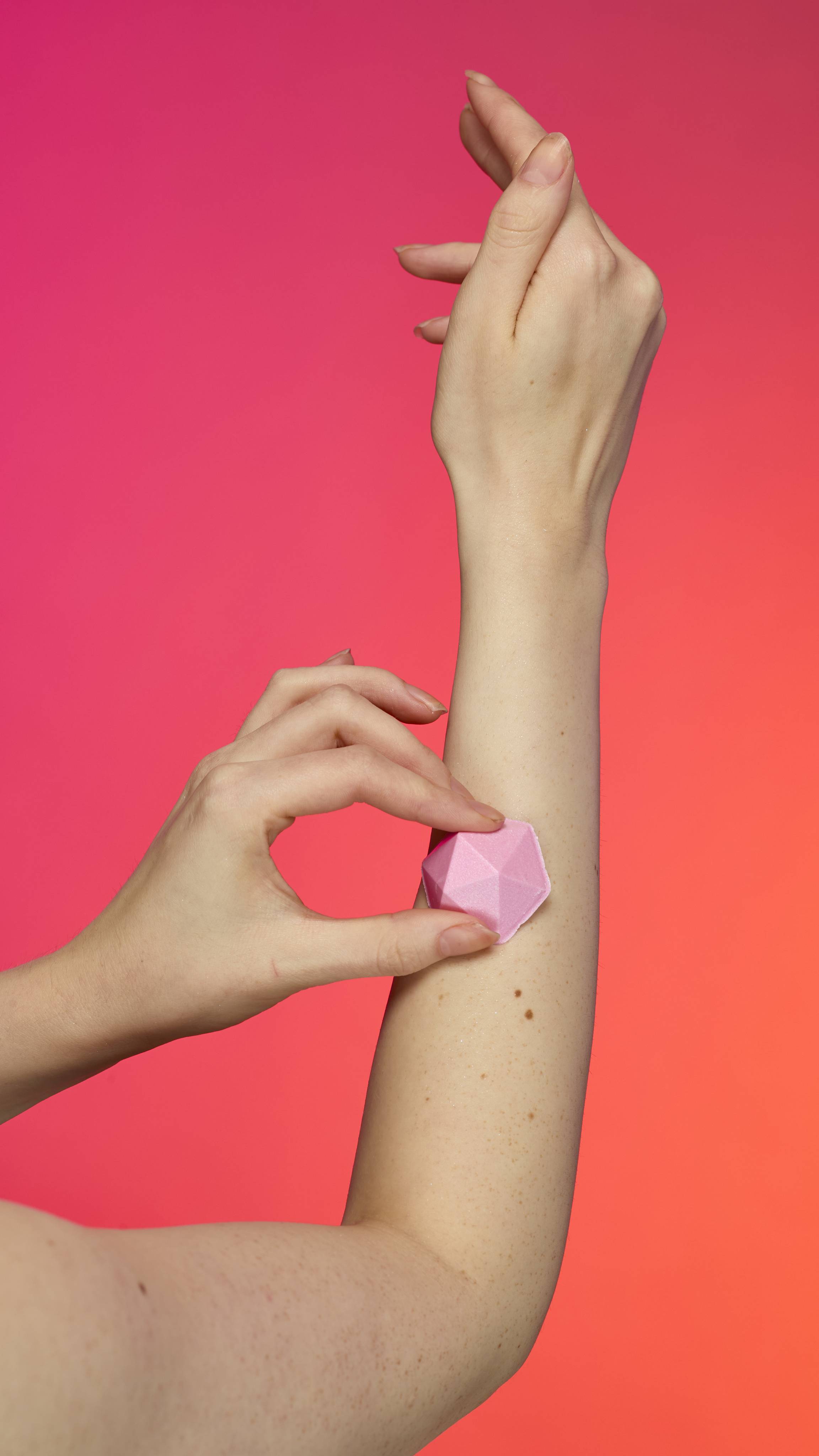 A person applies the pyramid-shaped Pink Glitterbug Body bar to their arm. The fair skin shimmers with plastic free glitter against a vibrant magenta and orange background.