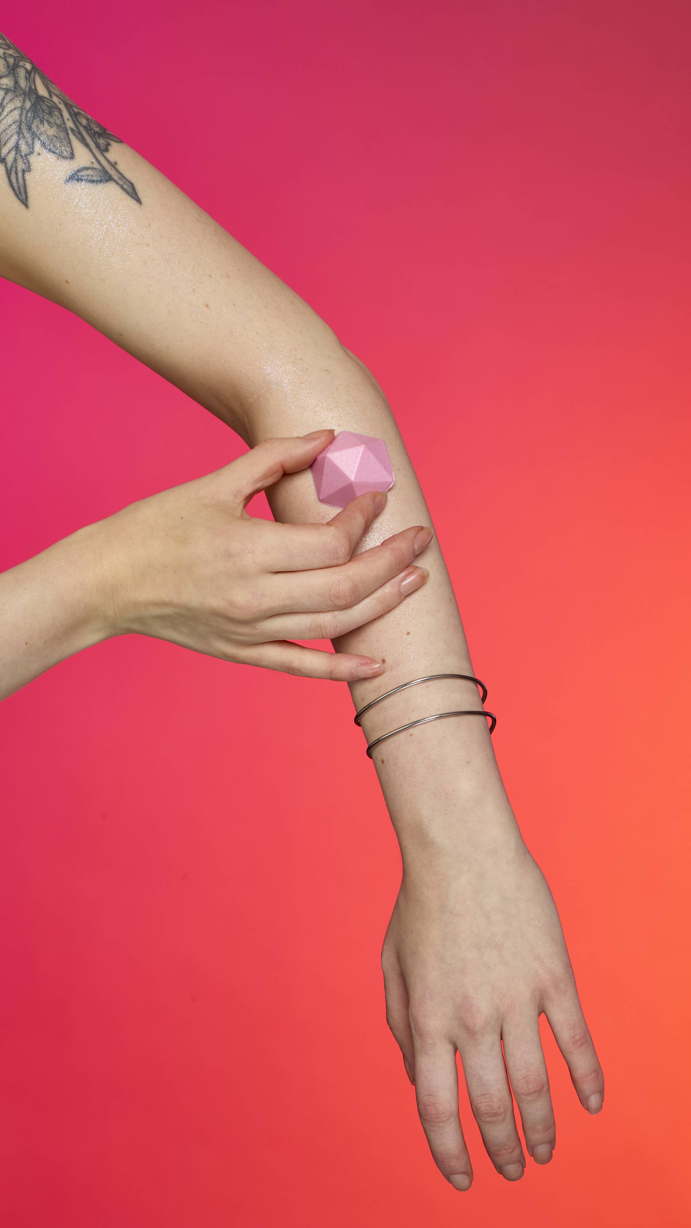 Two hands with fair skin apply the faceted, pyramid-shaped Pink Glitterbug Body bar to a tattooed arm. The arm has two thin bracelets and shimmers with plastic free glitter against a vibrant magenta and orange background.
